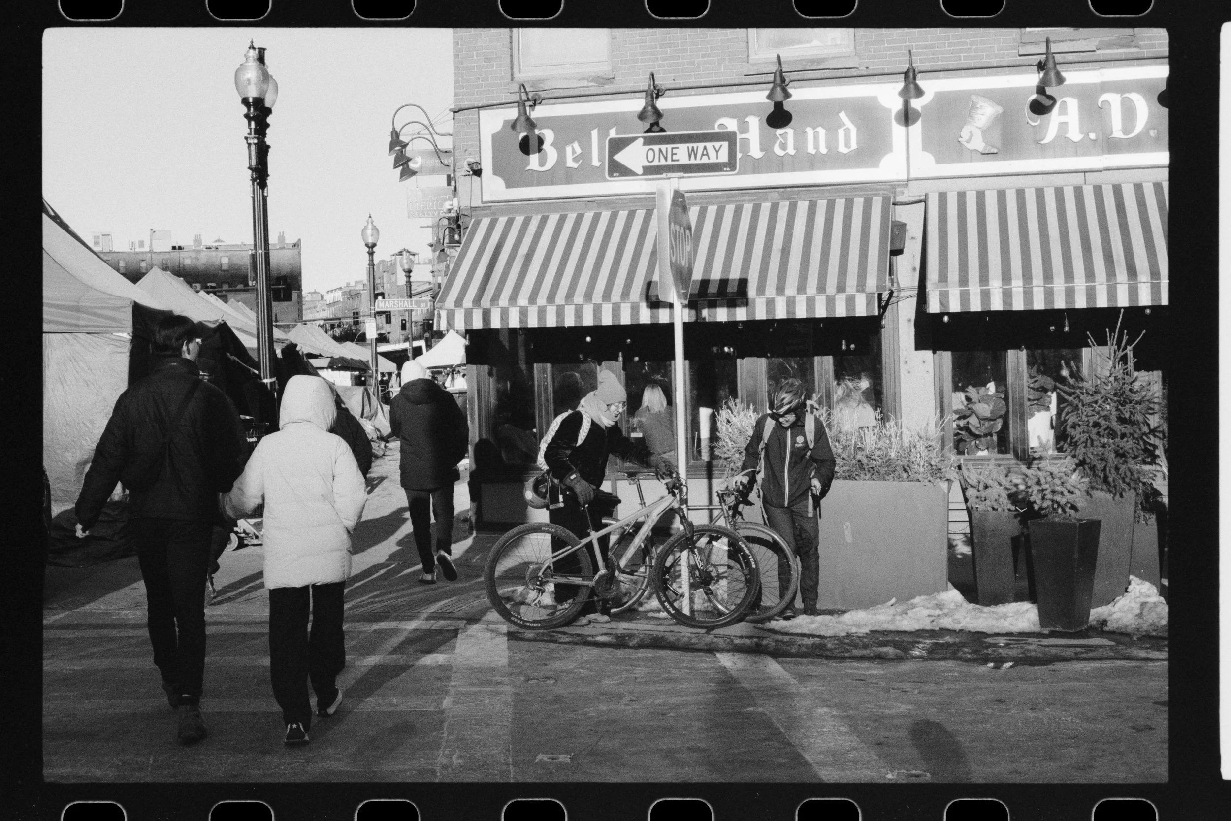 A black and white photo of a busy street scene with pedestrians, bicycles, and storefronts, including a store with striped awnings and a sign that reads 'Belle H...and A.D.'.
