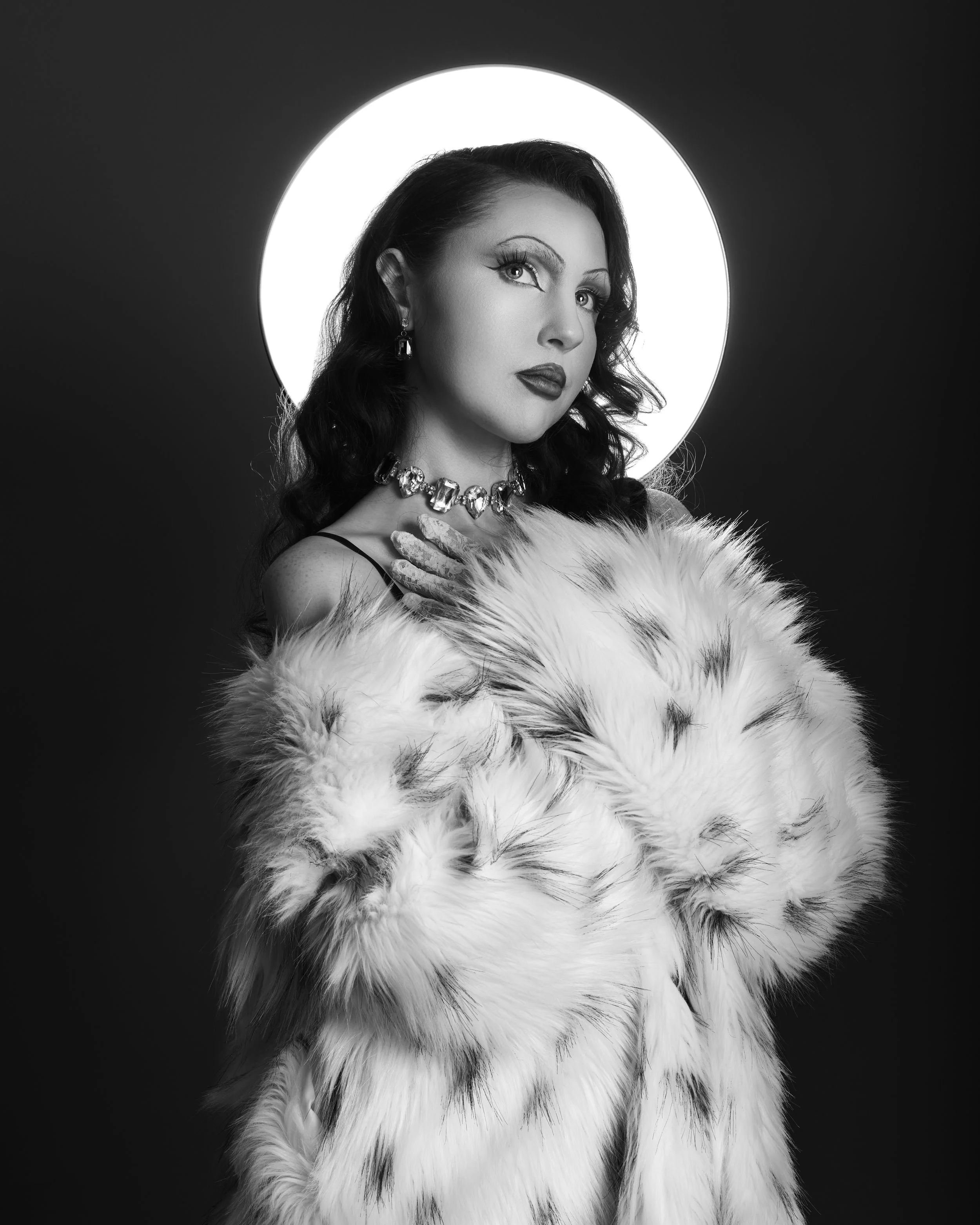 Black and white photo of a woman with dark, wavy hair, wearing a fur coat and jewelry, standing in front of a glowing circular light.