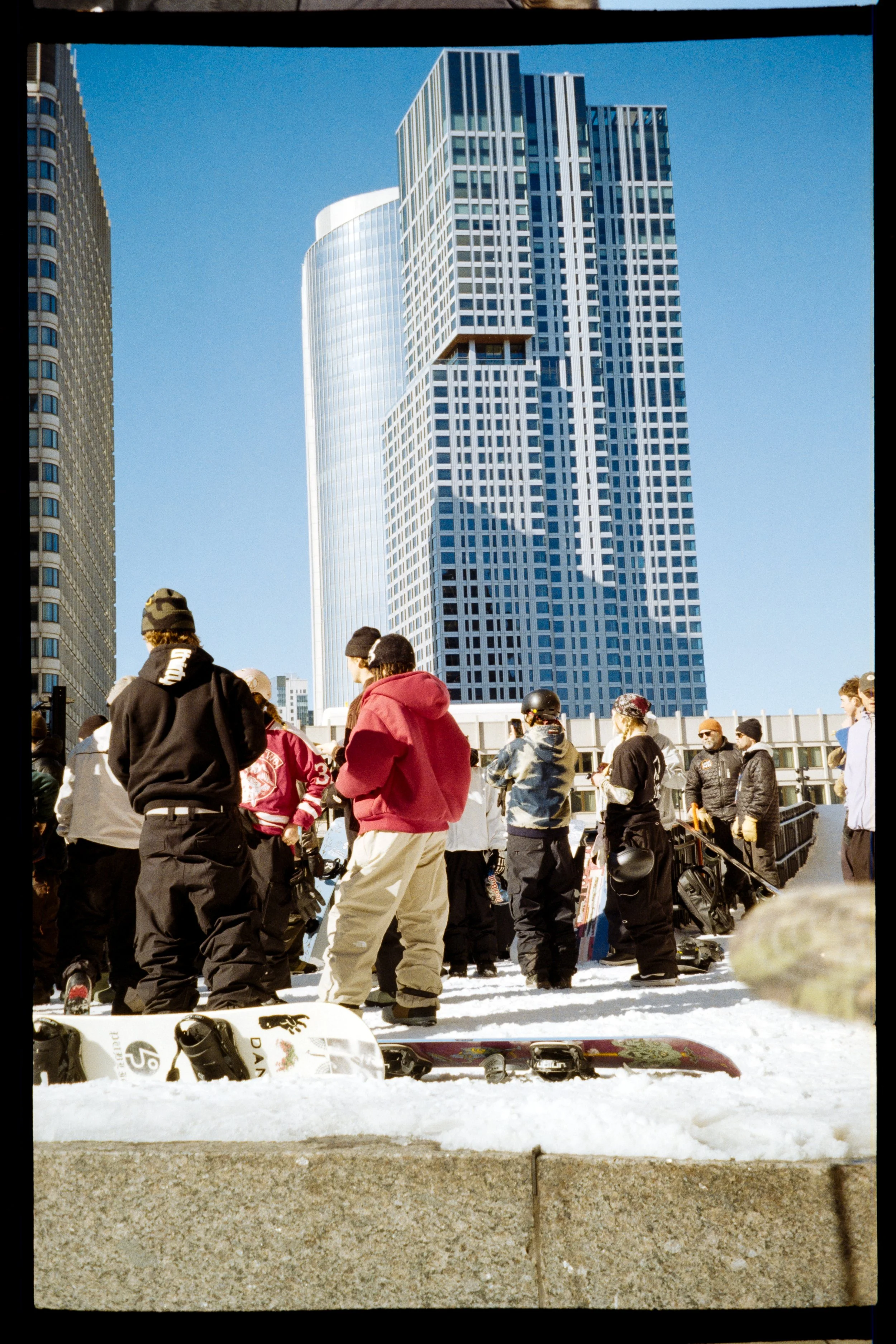 A group of people in winter clothing gathered outdoors on snow, with snowboard equipment at their feet and modern high-rise buildings in the background.
