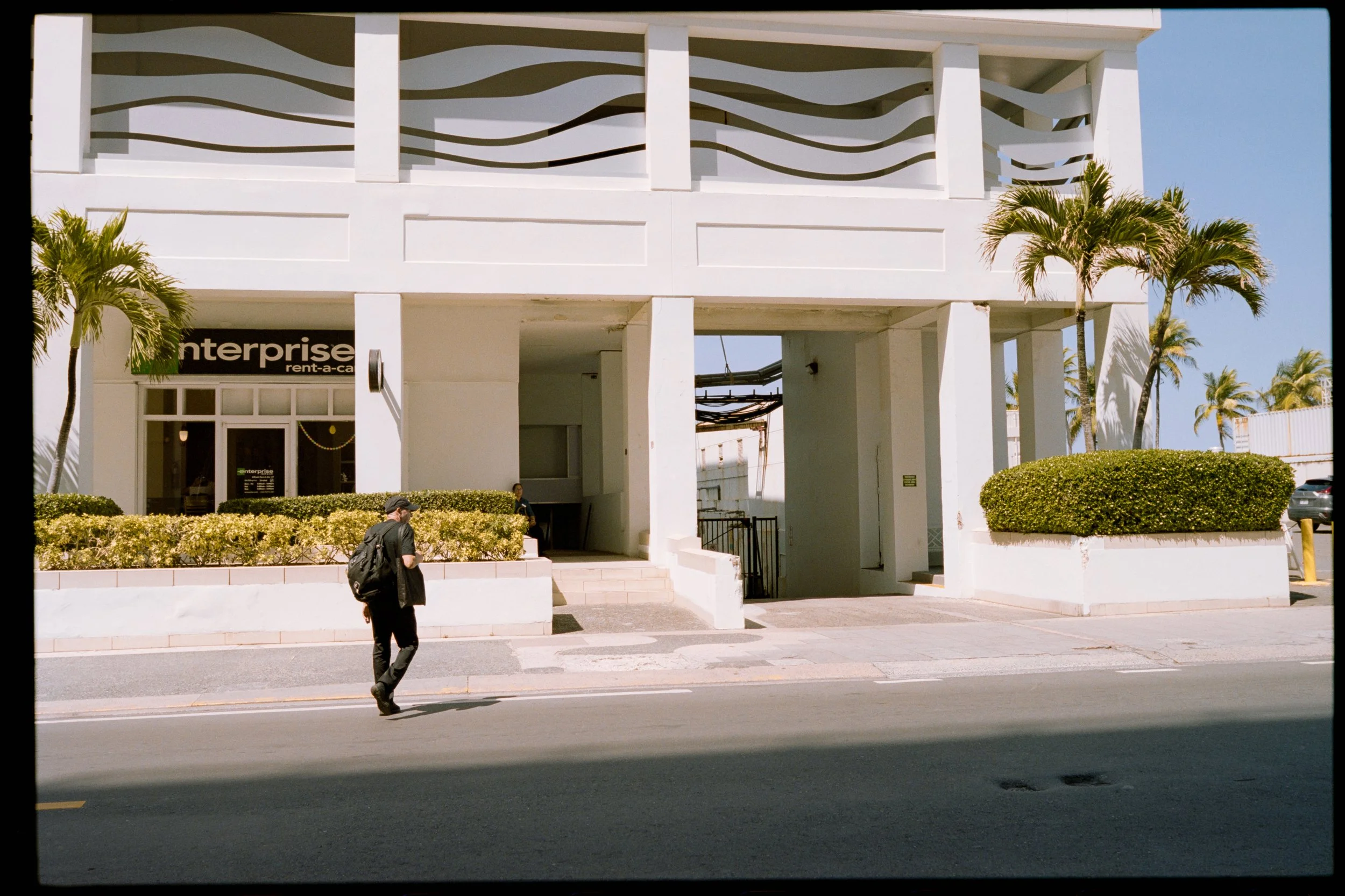 White multi-story building with decorative wave pattern on the balcony railing, palm trees in front, and a sign reading "enterprise rent-a-car" on the lower level. A person with a backpack is walking on the sidewalk in front of the building.