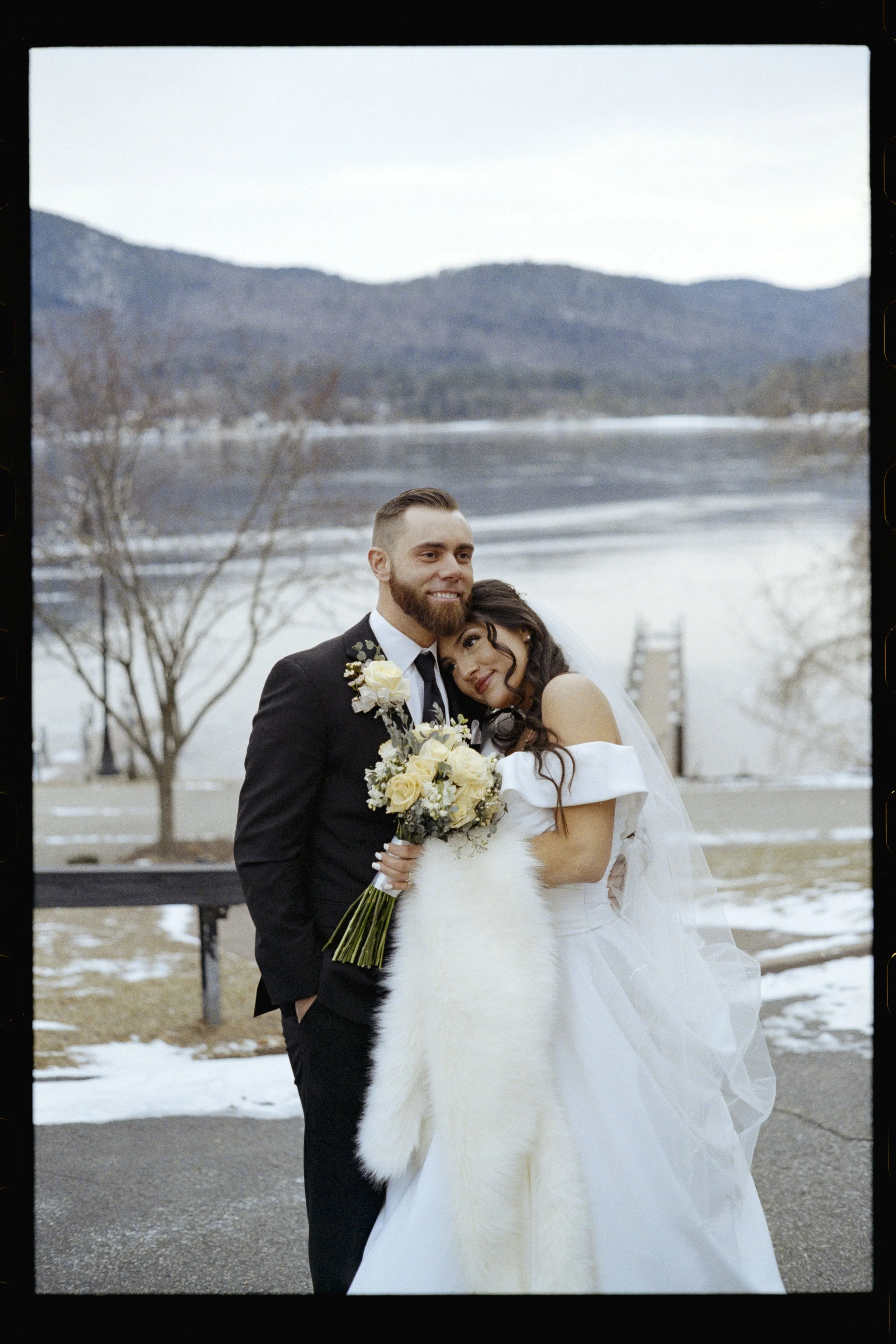 A bride and groom pose together outdoors near a lake with mountains in the background, on a cold day with patches of snow on the ground.
