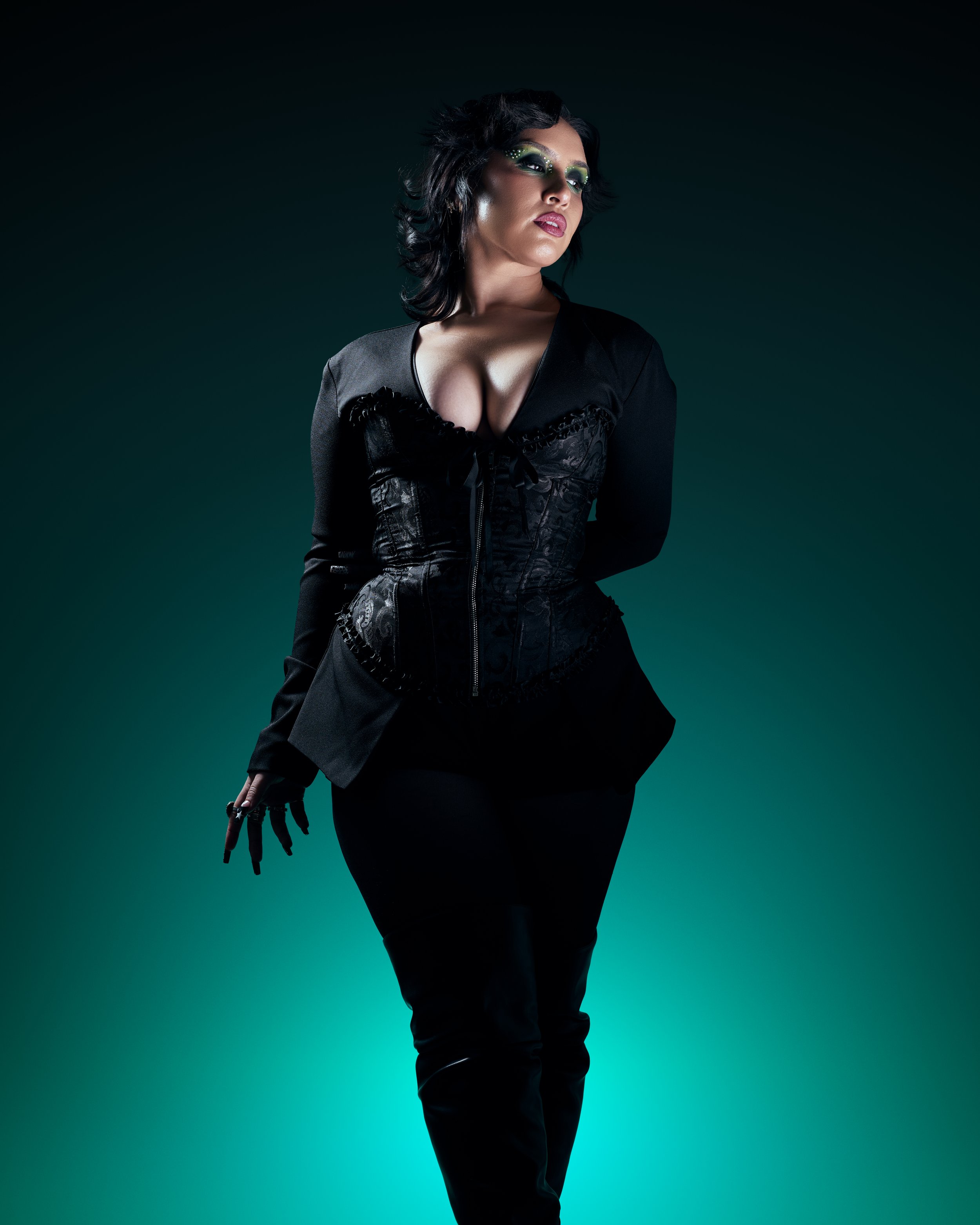 A woman with dark, wavy hair and makeup with green eyeshadow and dark lipstick, wearing a black lace corset, blazer, and pants, standing against a dark gradient background.