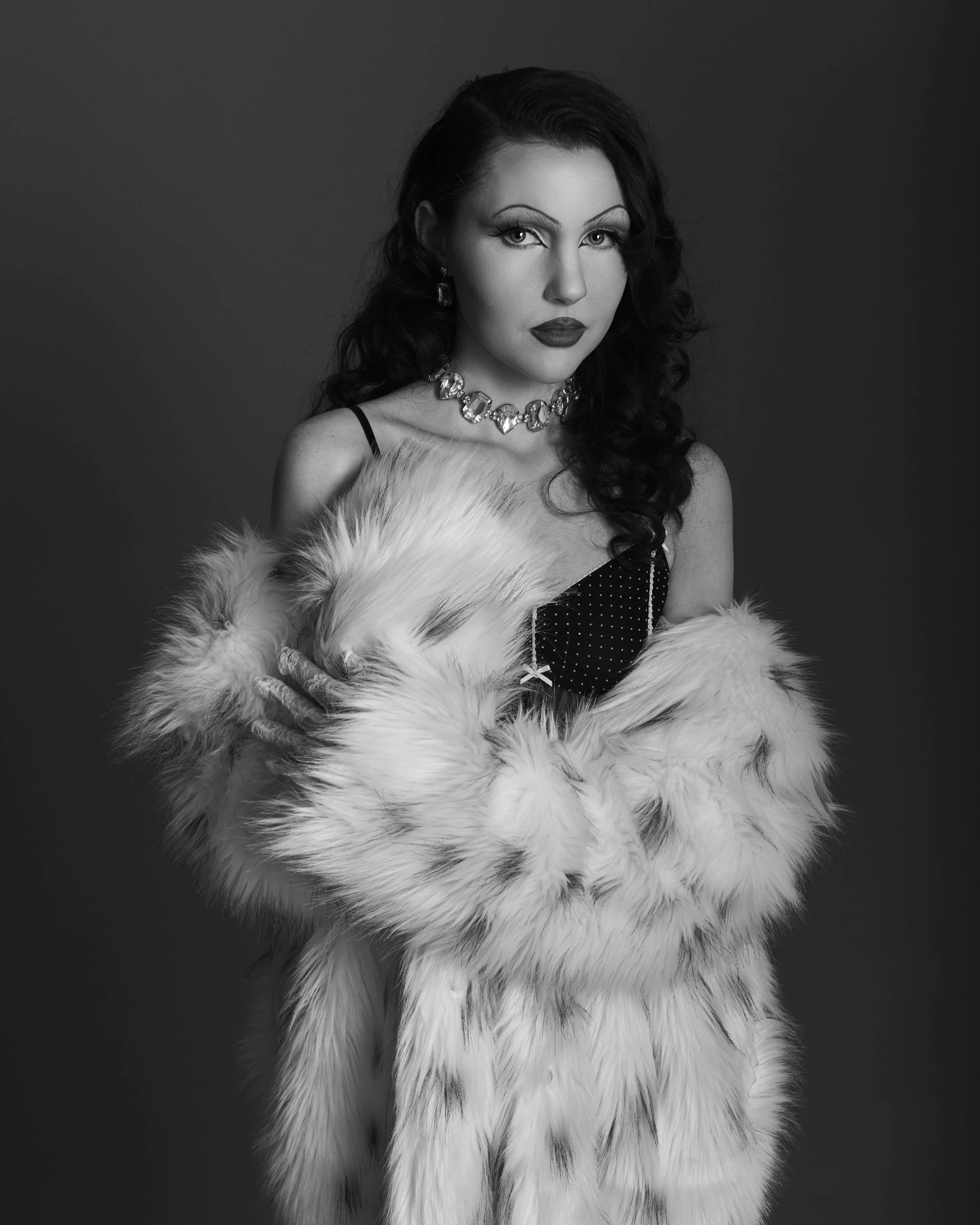 A black and white portrait of a woman with dark curly hair, wearing a fur coat, a necklace, and earrings, holding a small dog close to her chest.