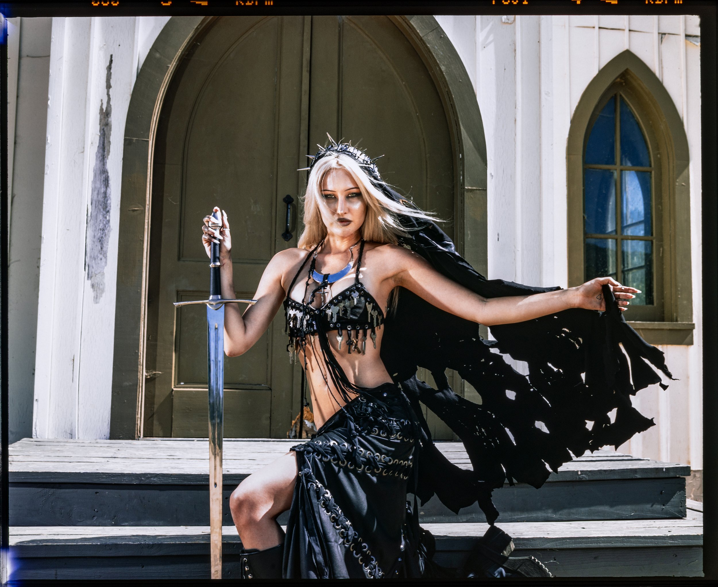 A woman dressed in black, wearing a crown of spikes, holding a sword, standing in front of a church with gothic windows and an arched door.