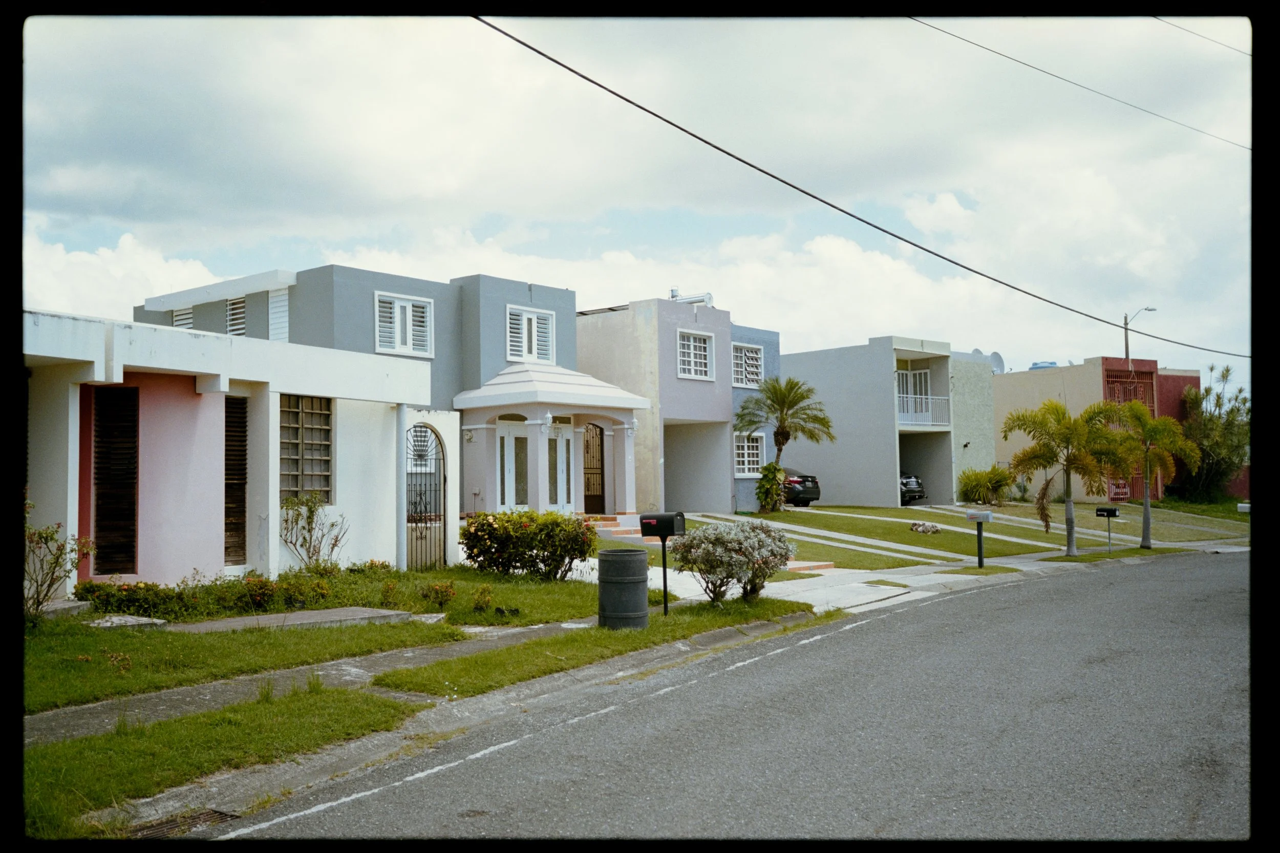 Row of modern residential houses with colorful facades, well-maintained front yards, palm trees, parked cars, and a sidewalk in a suburban neighborhood