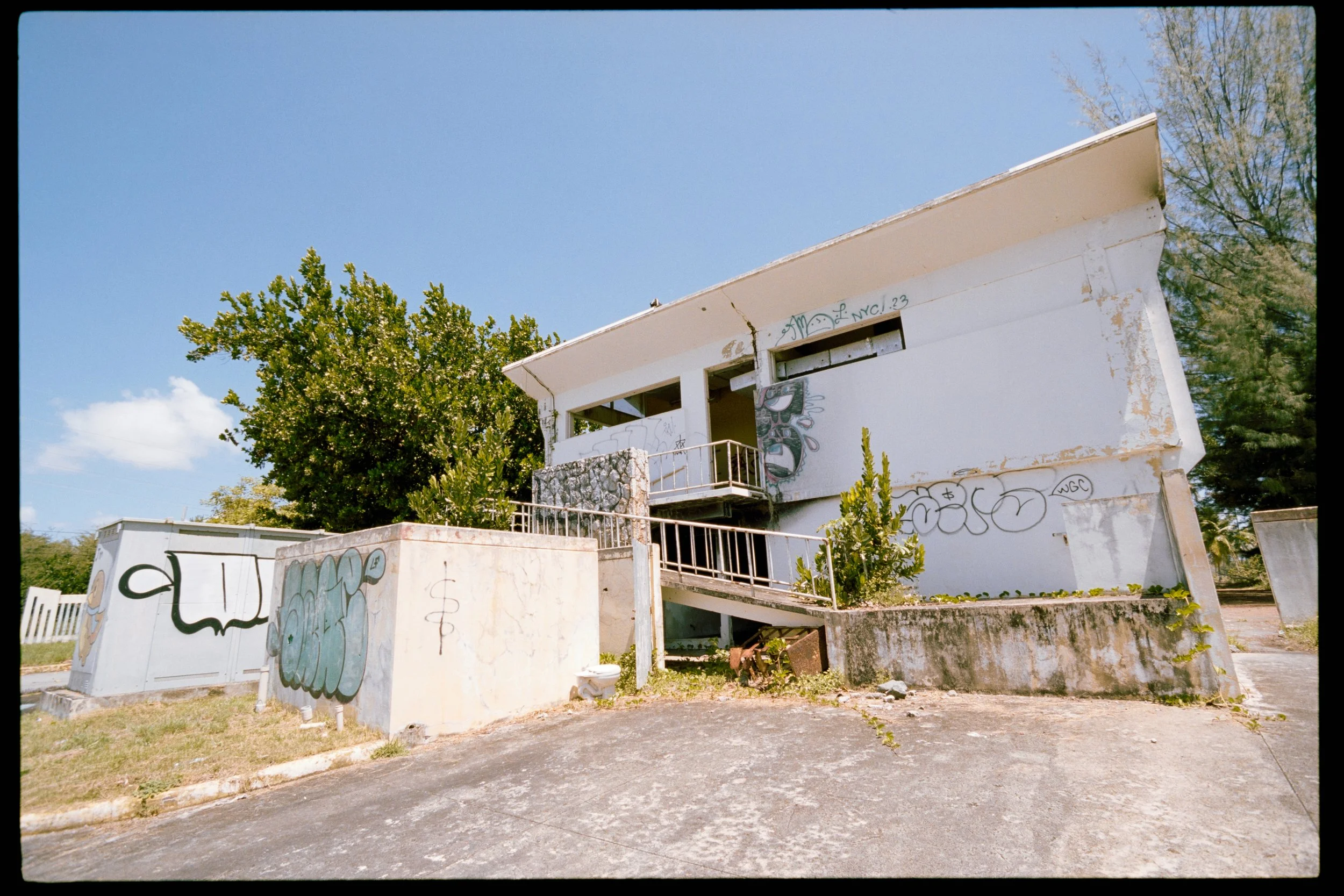 An abandoned building with graffiti on the walls, surrounded by trees and a cloudy blue sky.