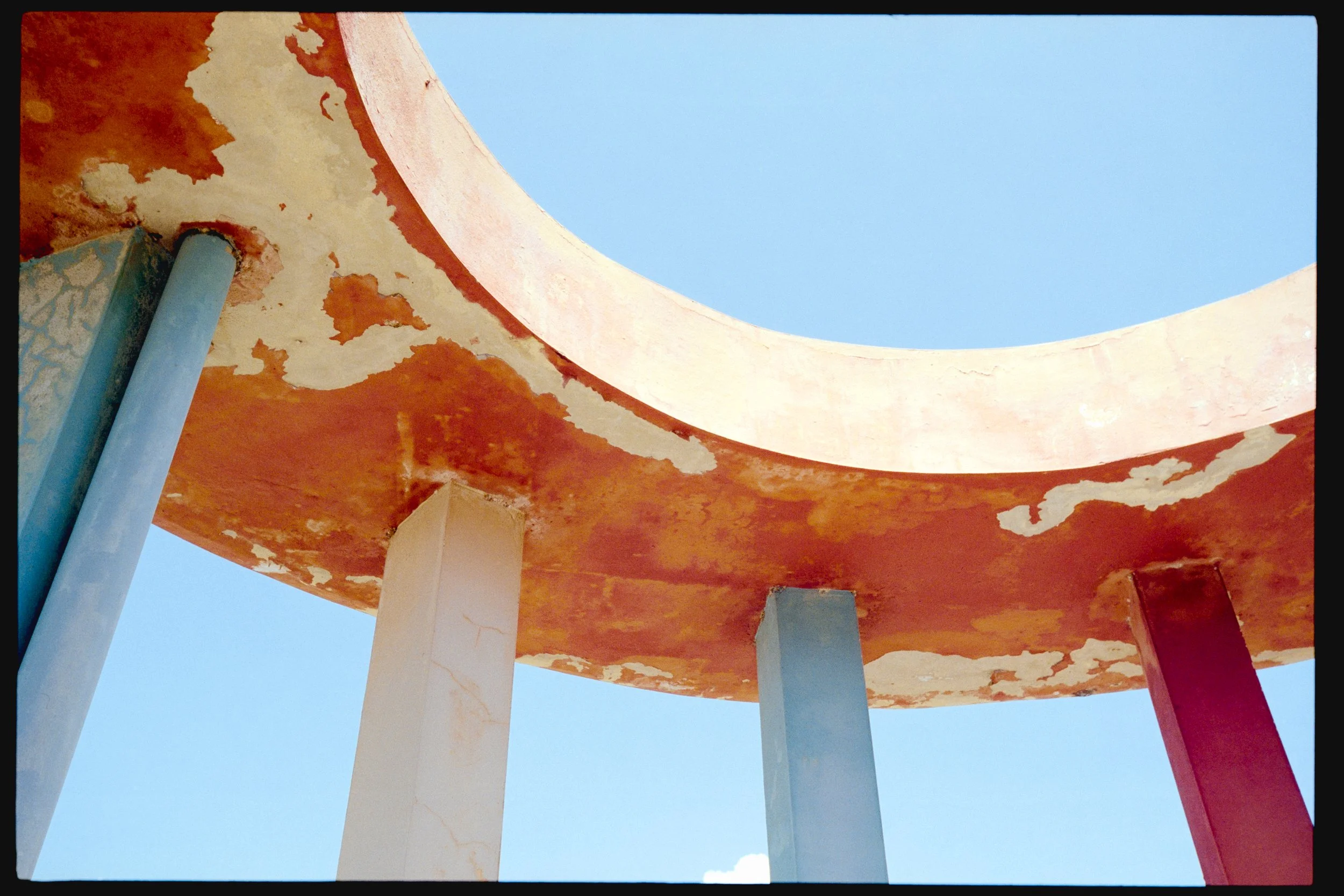 Looking up at a building with a curved, orange-colored roof supported by white columns, against a clear blue sky.