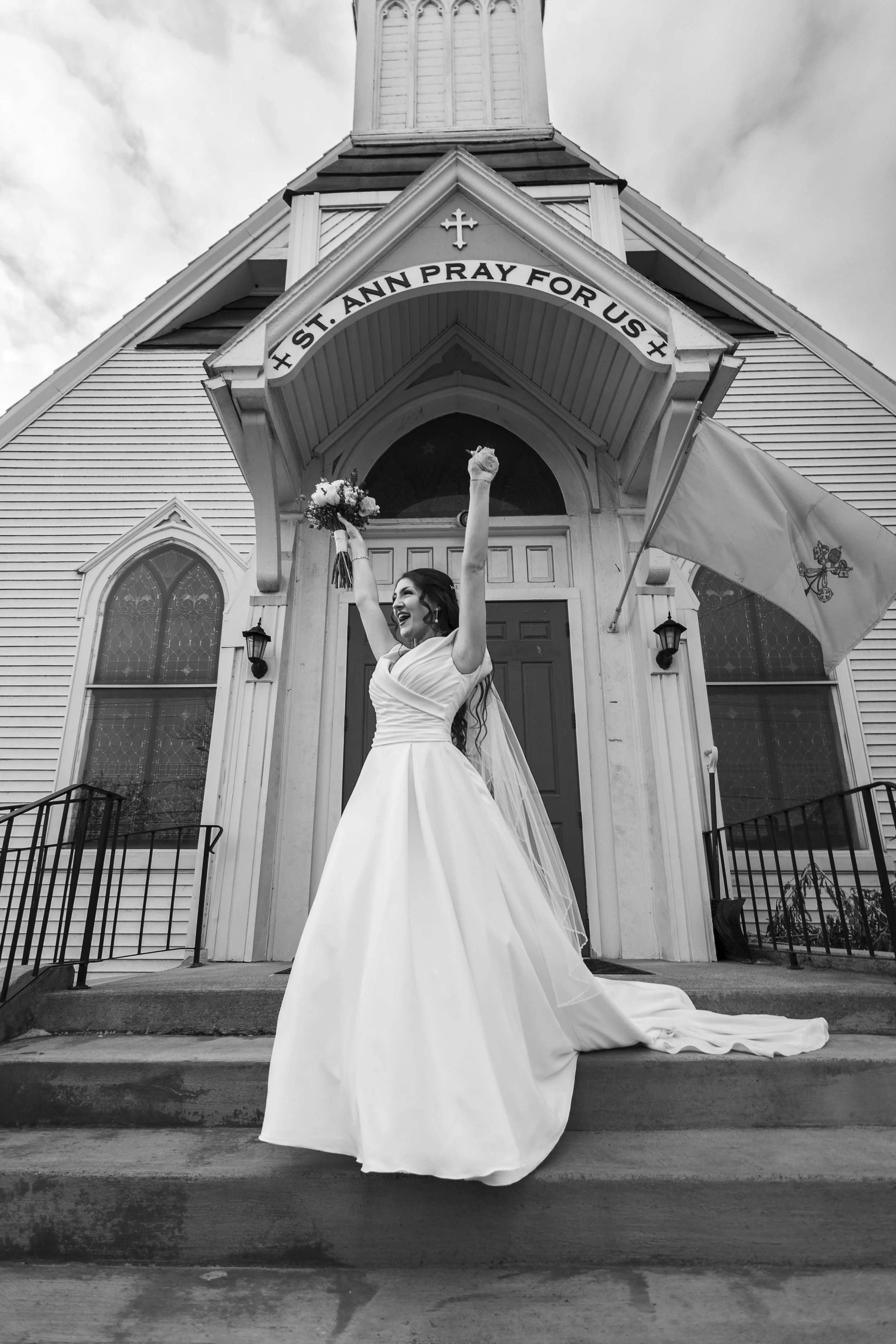 A bride in a wedding dress celebrating in front of a church with the sign "St. Ann Pray For Us."