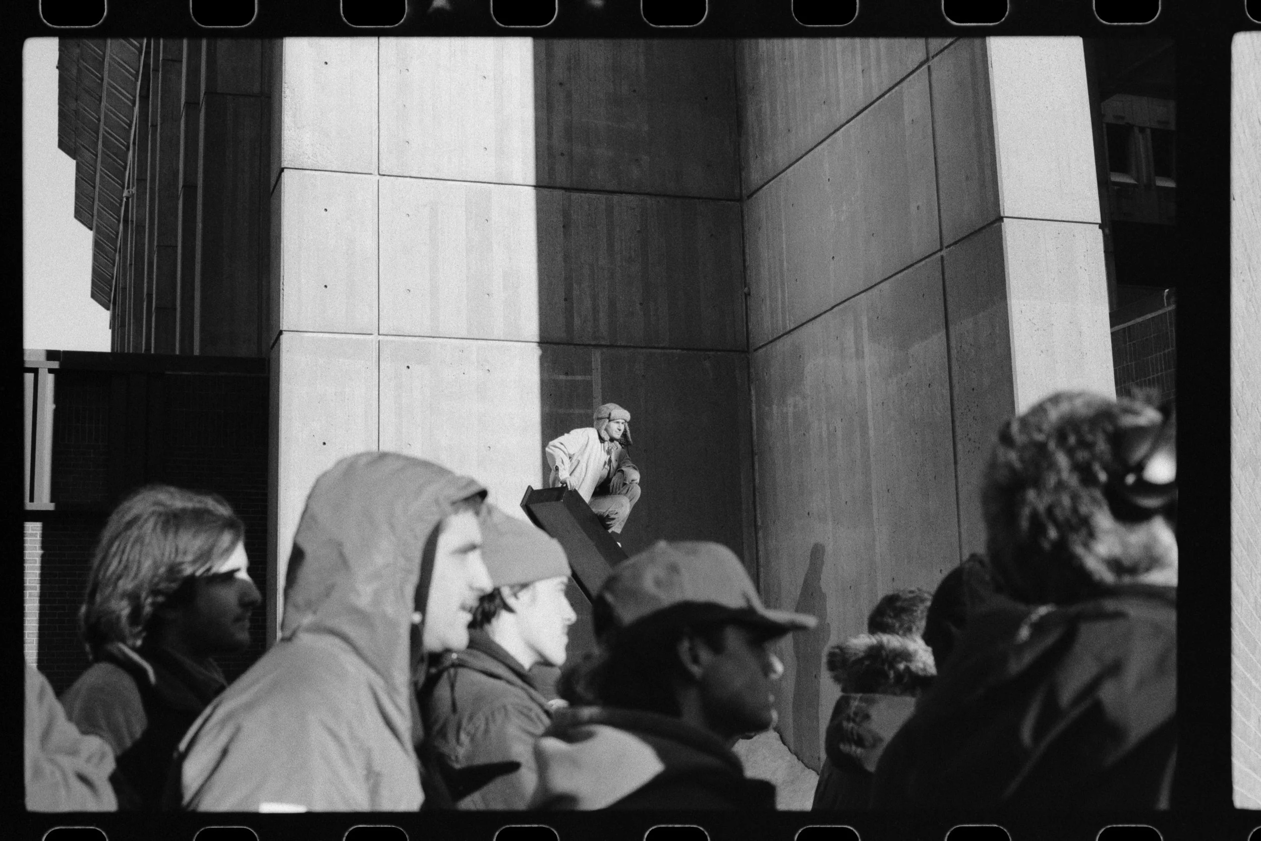 Black and white photo of a group of people, mostly young women, standing in line or crowd outdoors. In the background, a man is sitting on a ledge against a building wall, wearing a hat and jacket.