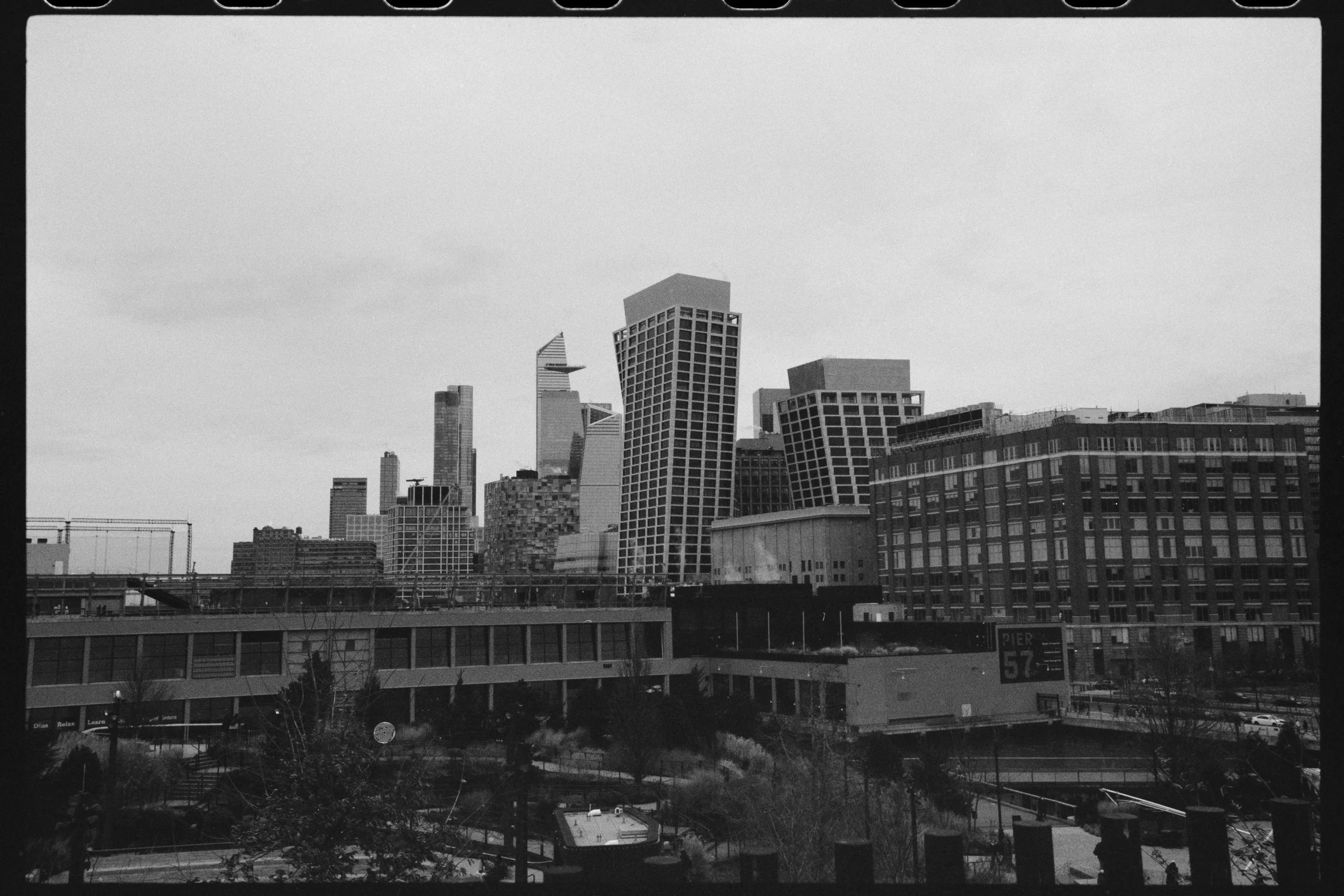 Black and white photograph of a city skyline with tall modern skyscrapers, some with unique architectural designs, seen from a lower vantage point. In the foreground, there is a multi-level city street and some trees.