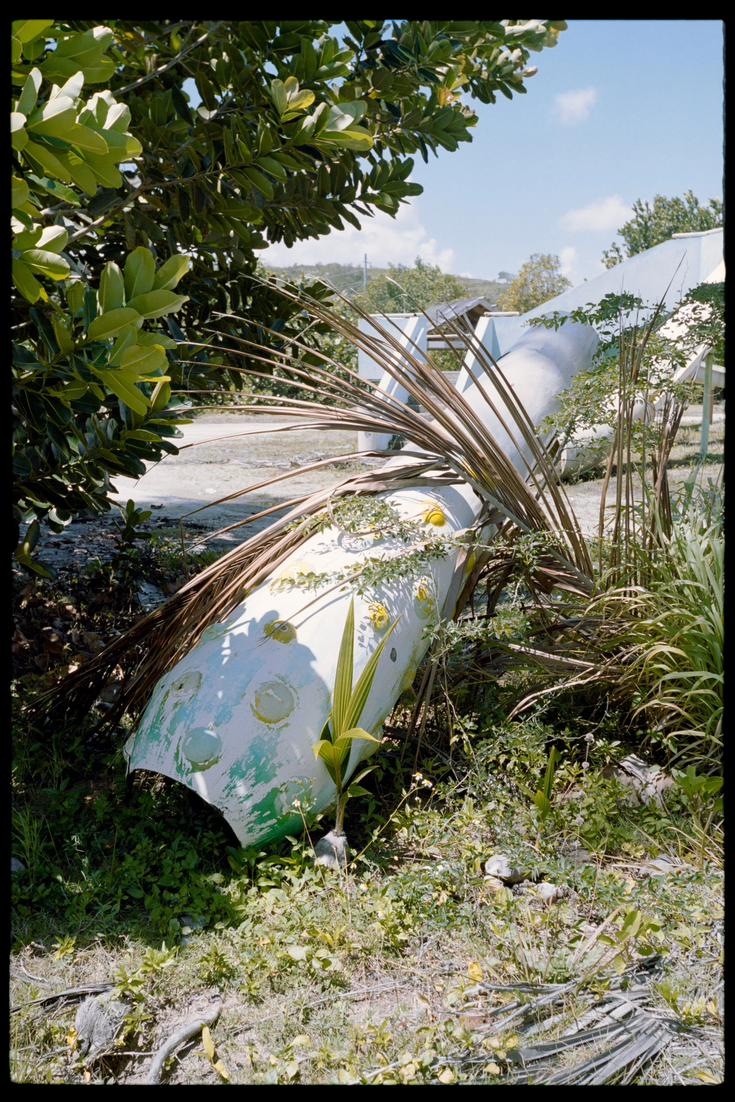 Collapsed large white pipe lying on the ground surrounded by green bushes, small plants, and dry grass in an outdoor area with a blue sky and some clouds in the background.