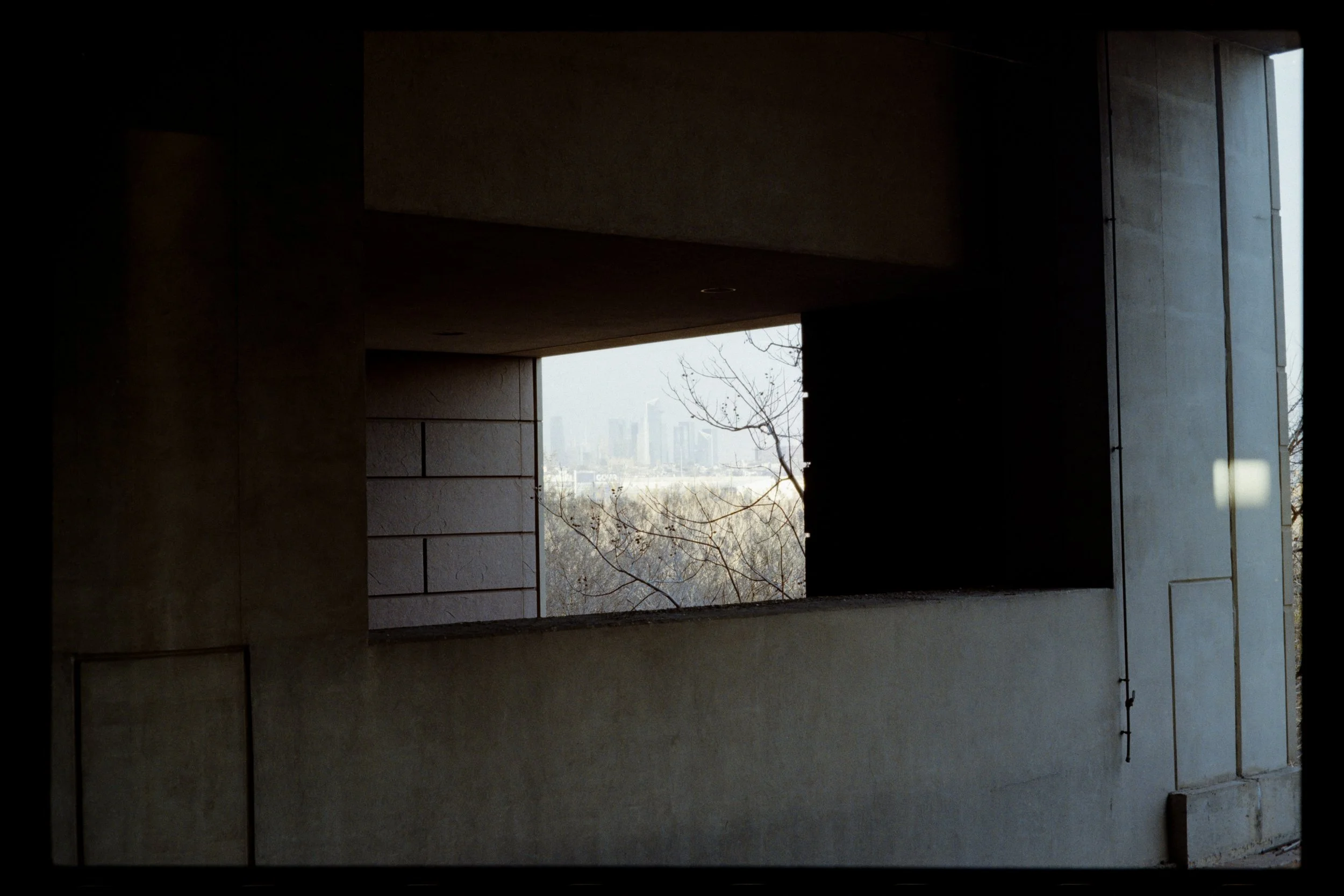 View through a concrete window opening of a city skyline with tall buildings, leafless tree branches, and a hazy sky.