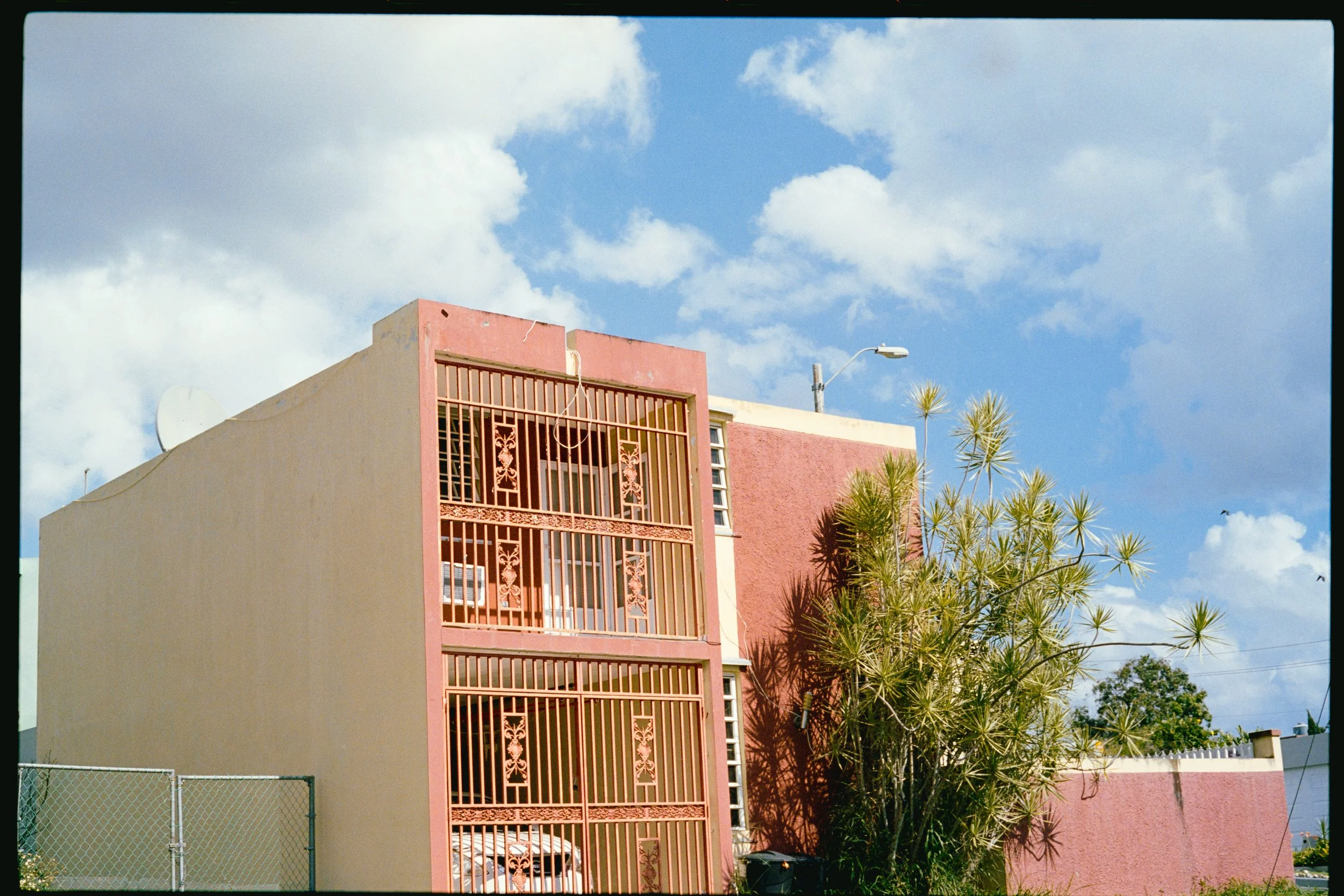 A building with pink and beige exterior walls, a decorative balcony with ironwork, a satellite dish on the roof, and a tree with long, thin leaves in front. The sky is blue with scattered white clouds.