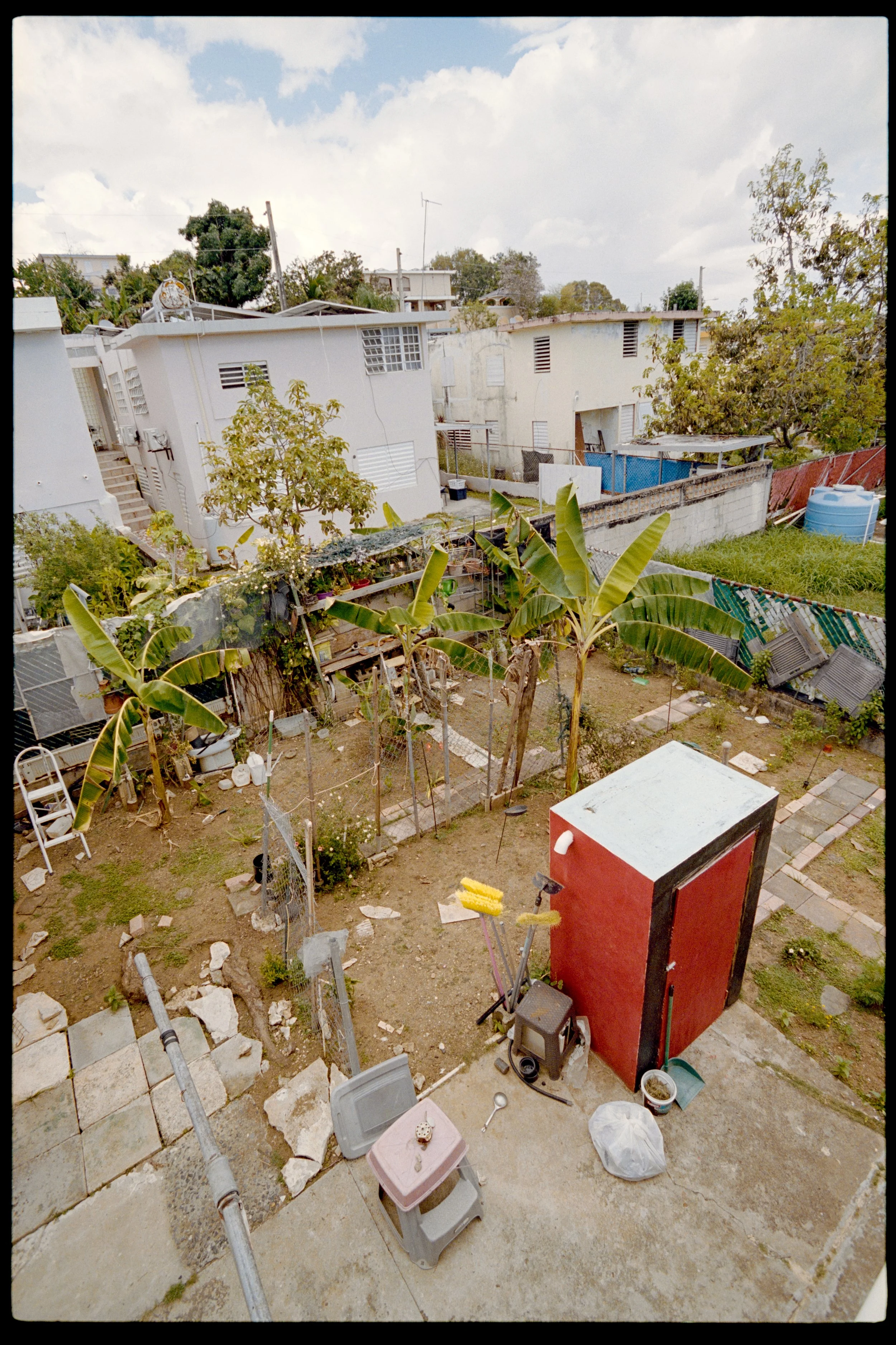 Backyard scene with laundry, miscellaneous items, plants, and surrounding houses under cloudy sky.