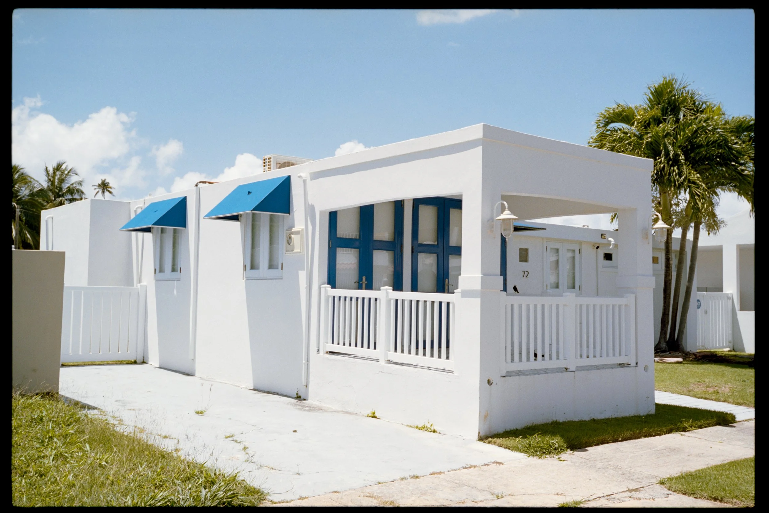 White house with blue awnings, blue door, white fence, palm trees, and a blue sky with clouds.