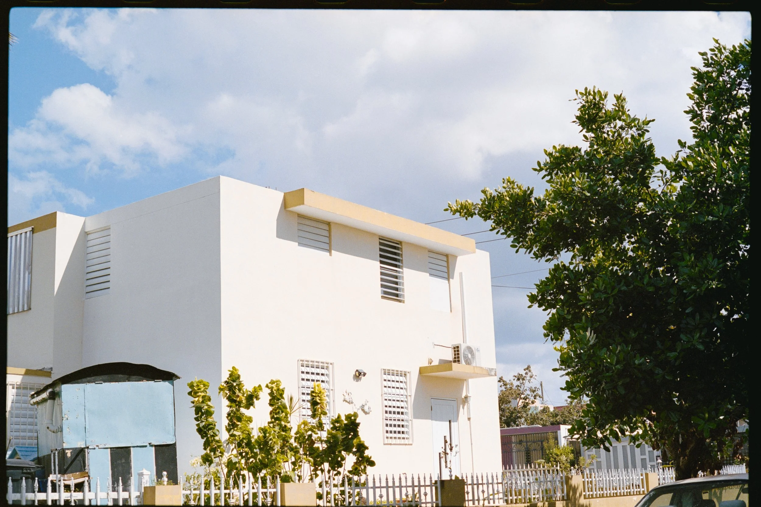 A white modern house with flat roof, several windows with horizontal slats, and a small balcony. There is greenery including trees and plants around the house, with a clear blue sky and some clouds overhead.