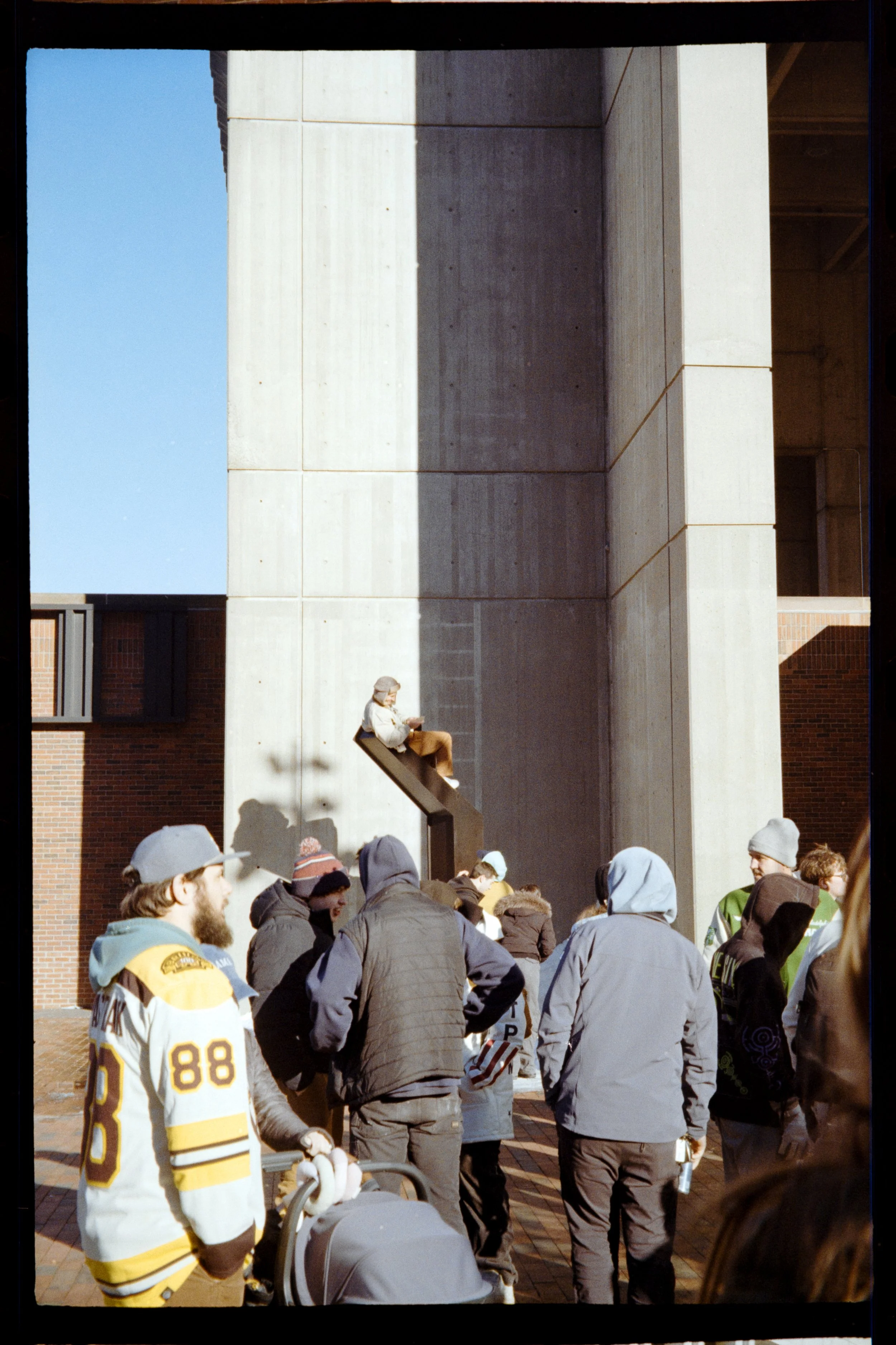 A crowd of people gathered outside a building with a large concrete wall. One person is sitting on a ledge, and others are standing and walking around, dressed in winter clothing and hats.