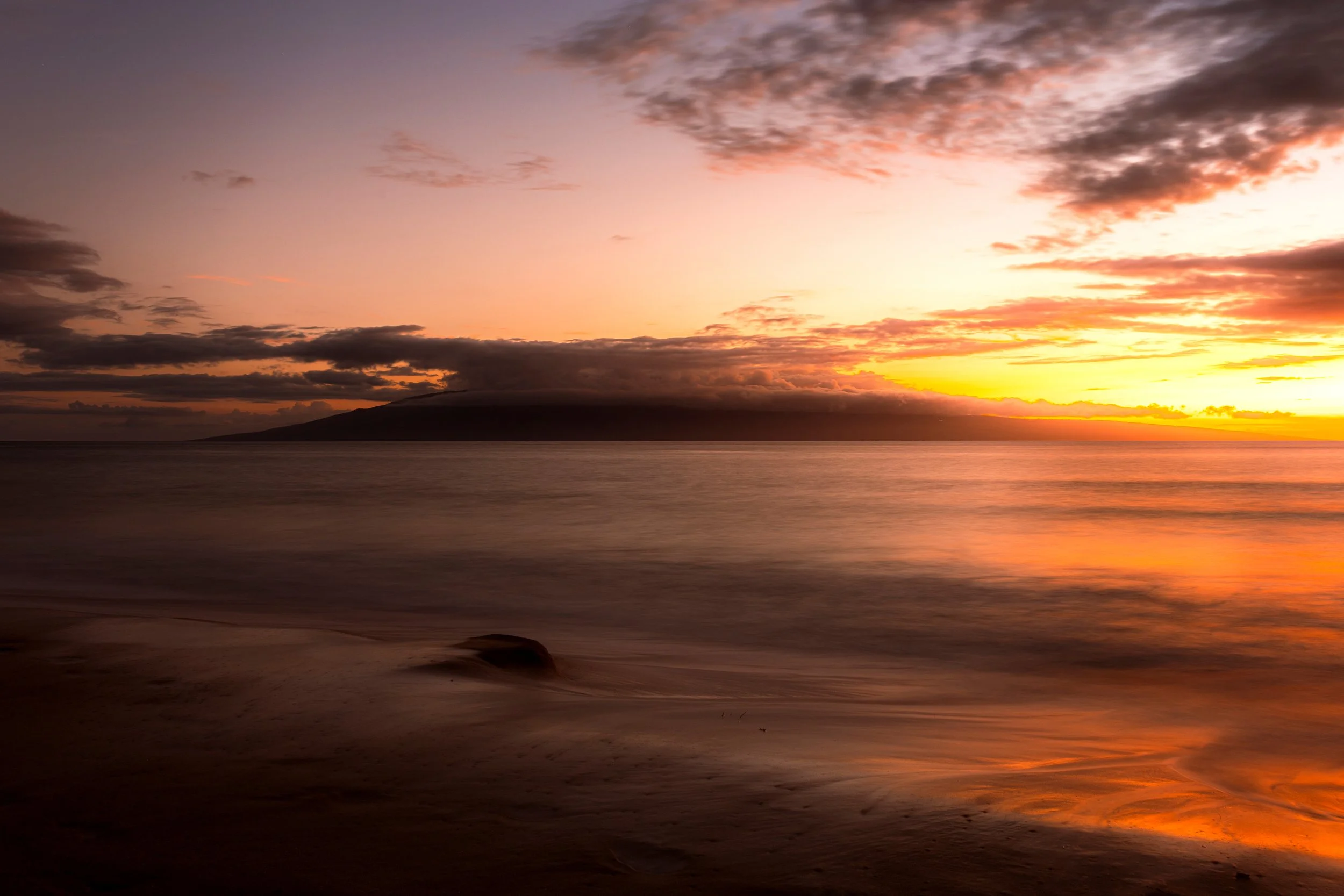 A scenic sunset over the ocean with a distant island silhouette, colorful sky with pink, orange, and purple clouds, and calm water reflecting the sunset colors.
