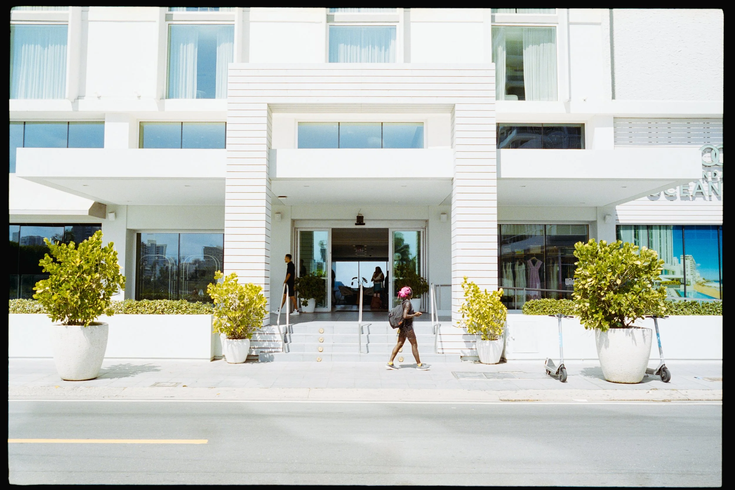 Front view of a modern white building with large windows and potted plants outside. People are walking into and out of the building. There are scooters parked on the sidewalk.