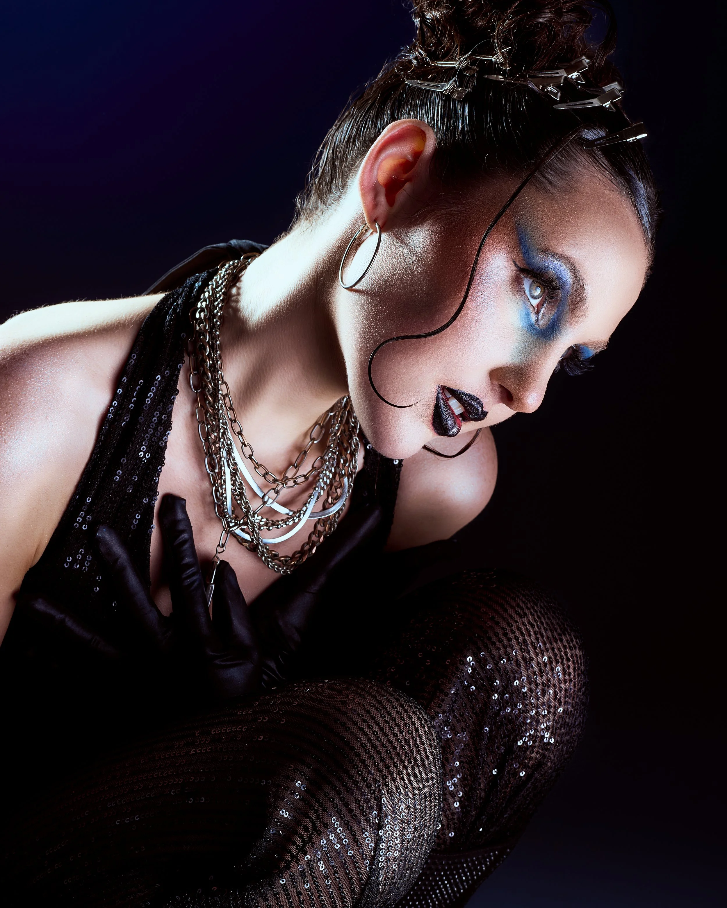 A woman in dark clothing with layered necklaces, earrings, black gloves, and sequin tights, exhibits a dramatic pose with makeup featuring blue eyeshadow and black lipstick.