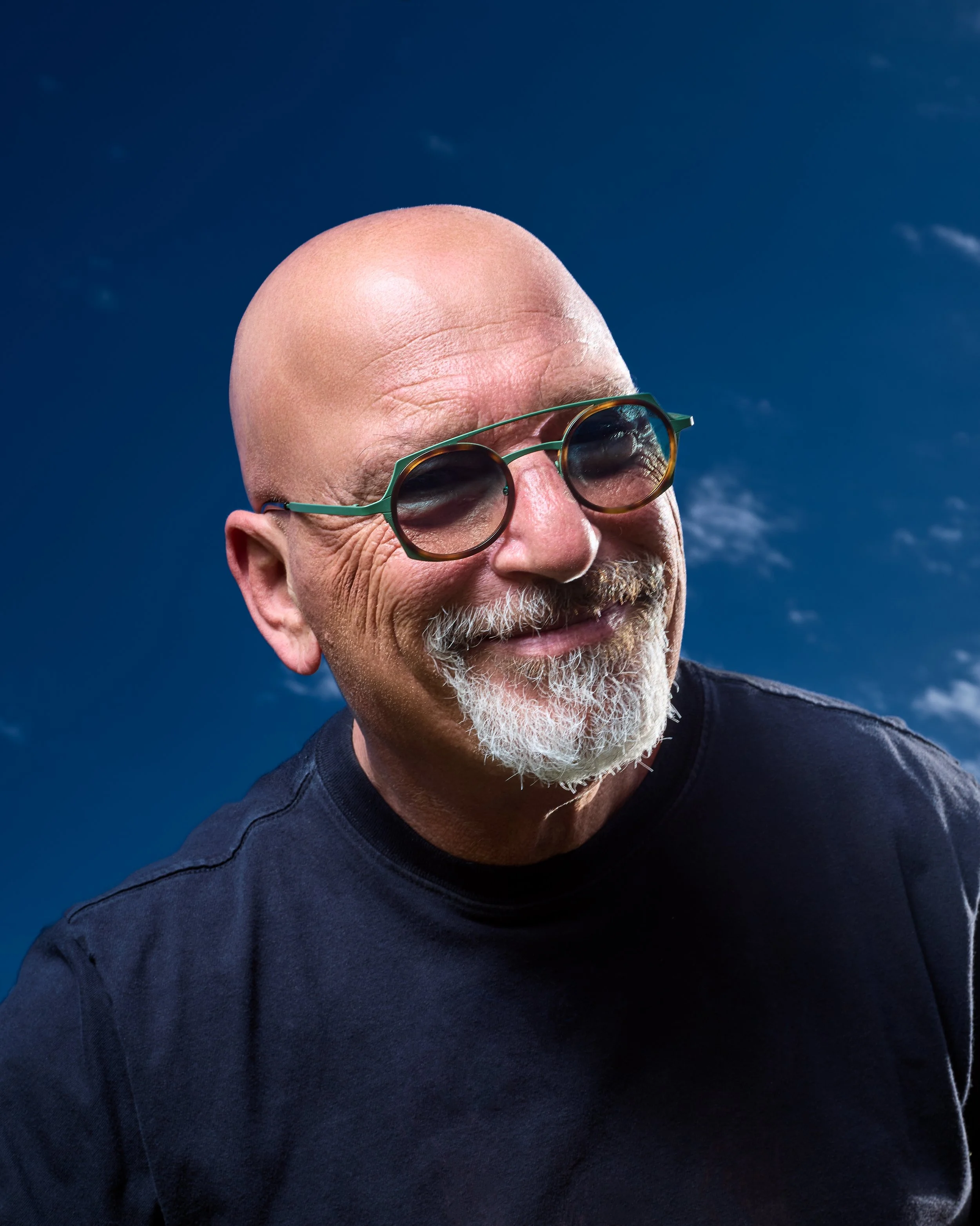 Smiling older man with a bald head, gray beard and mustache, wearing dark sunglasses and a black T-shirt against a background of a blue sky with some clouds.