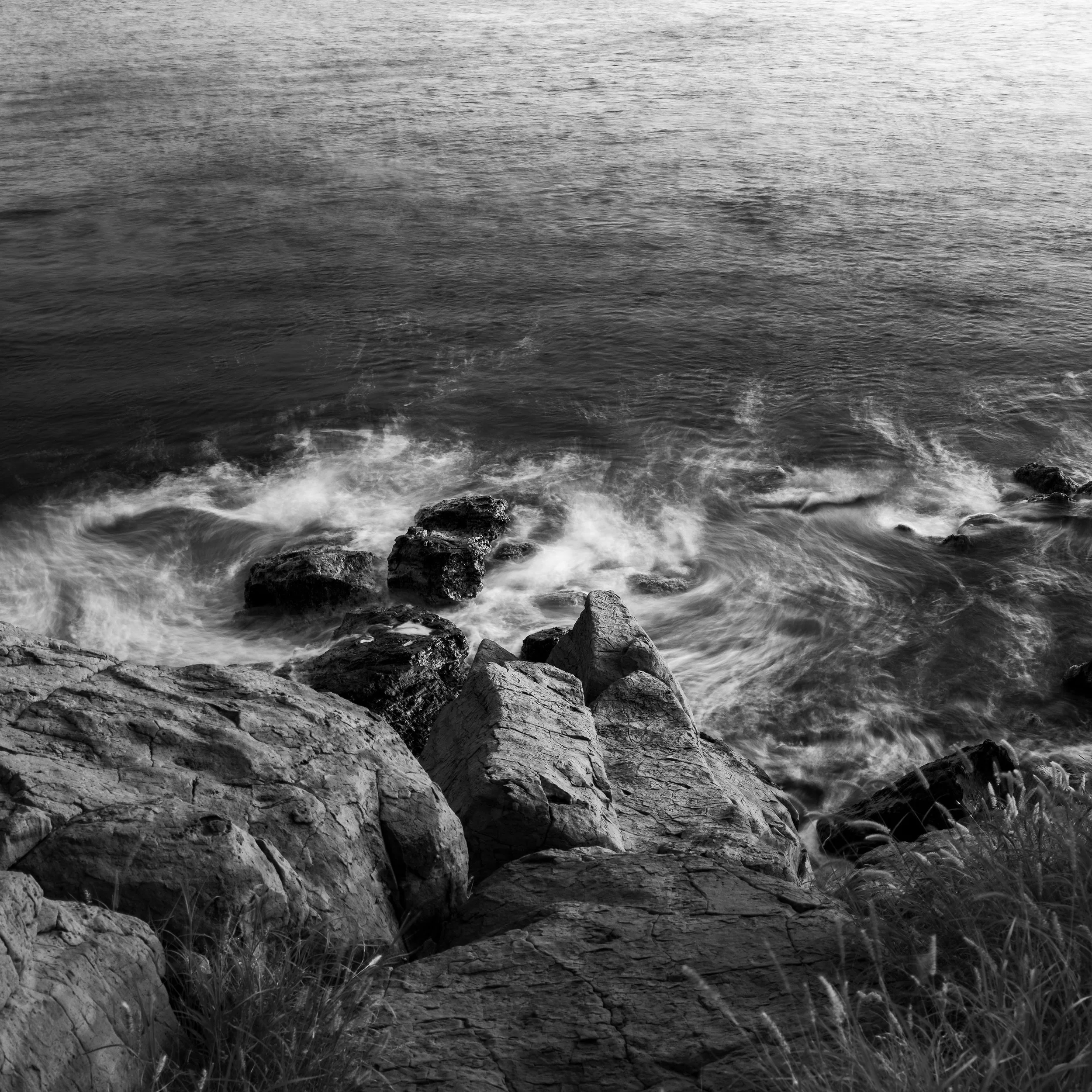 Black and white photograph of rocks along a shoreline, with water lapping against them and some rocks partially submerged.