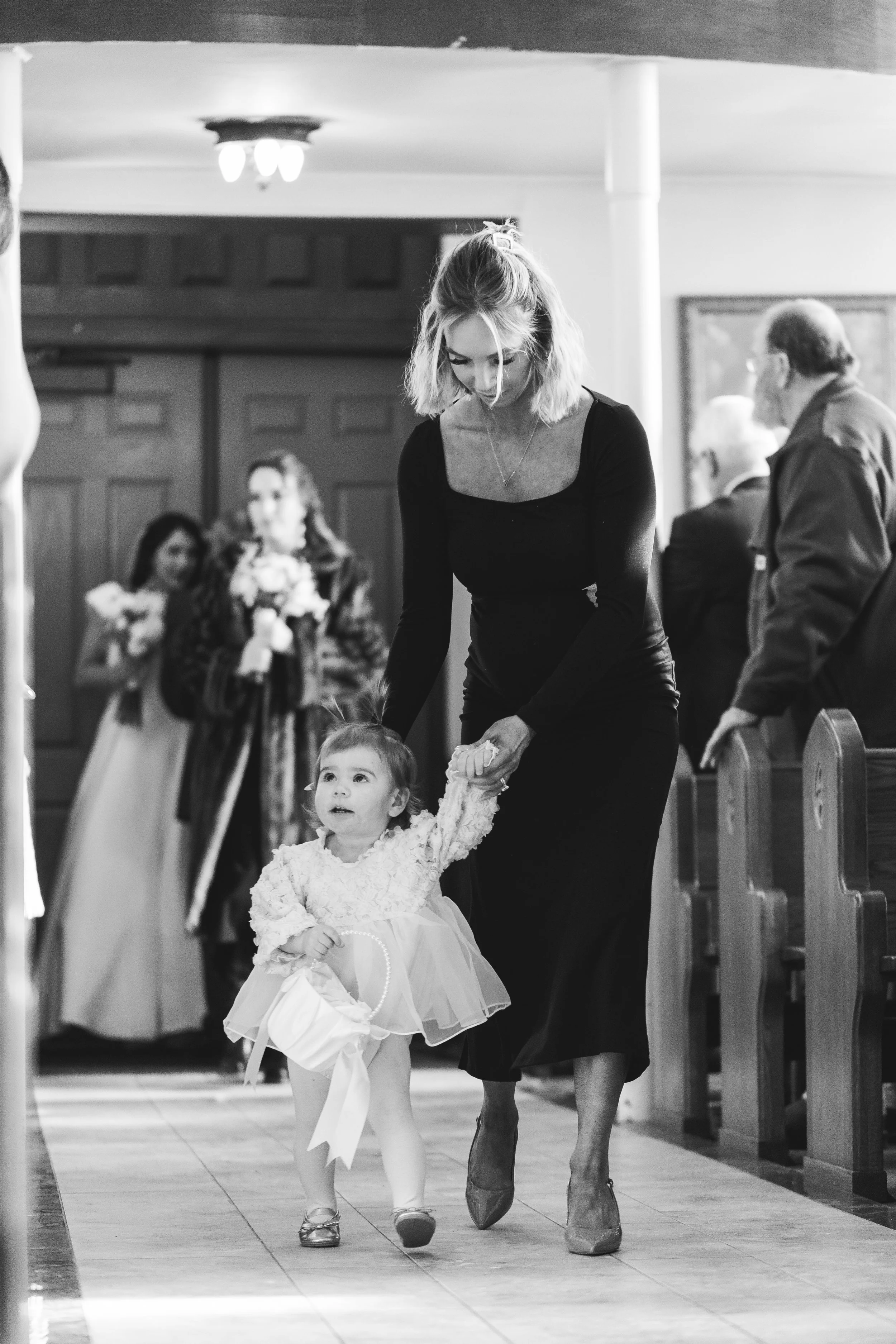 A woman helping a young girl down the aisle during a wedding in a church, with guests and a bride holding flowers in the background.