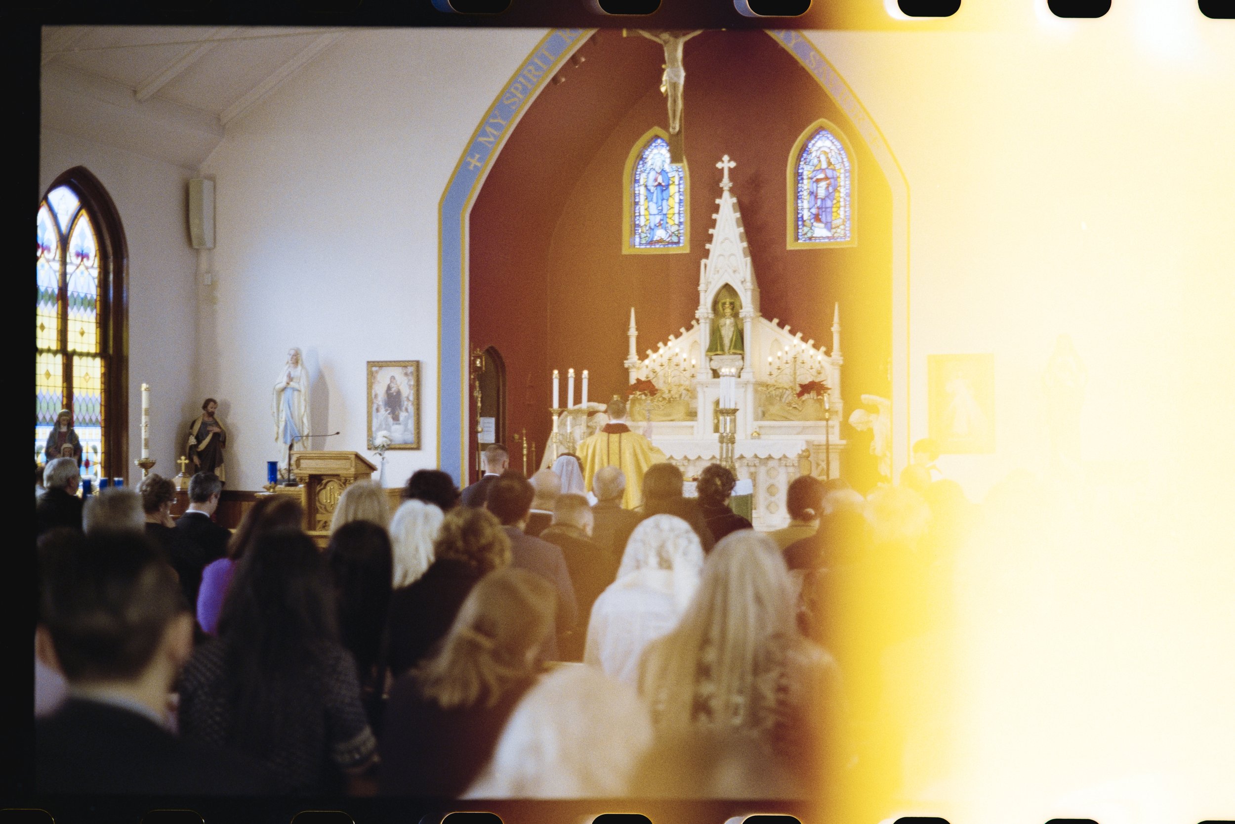 A congregation of people inside a church during a religious ceremony. The altar features a crucifix and stained glass windows, with clergy conducting the service.