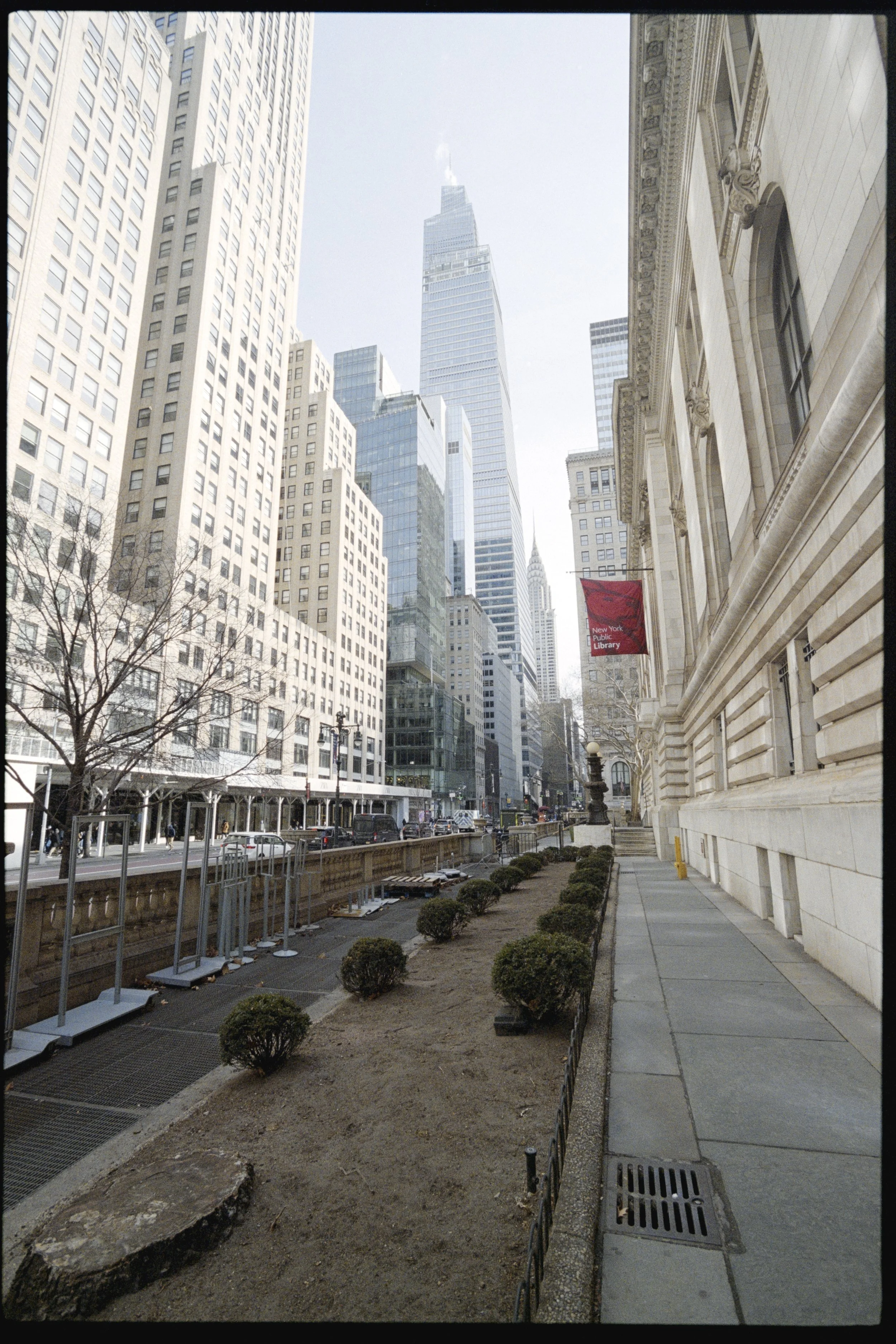 City street view with tall buildings, including the Empire State Building in the background. Sidewalk and a small landscaped area with bushes and soil, some construction elements, and a historic building with ornate architecture on the right. A red b