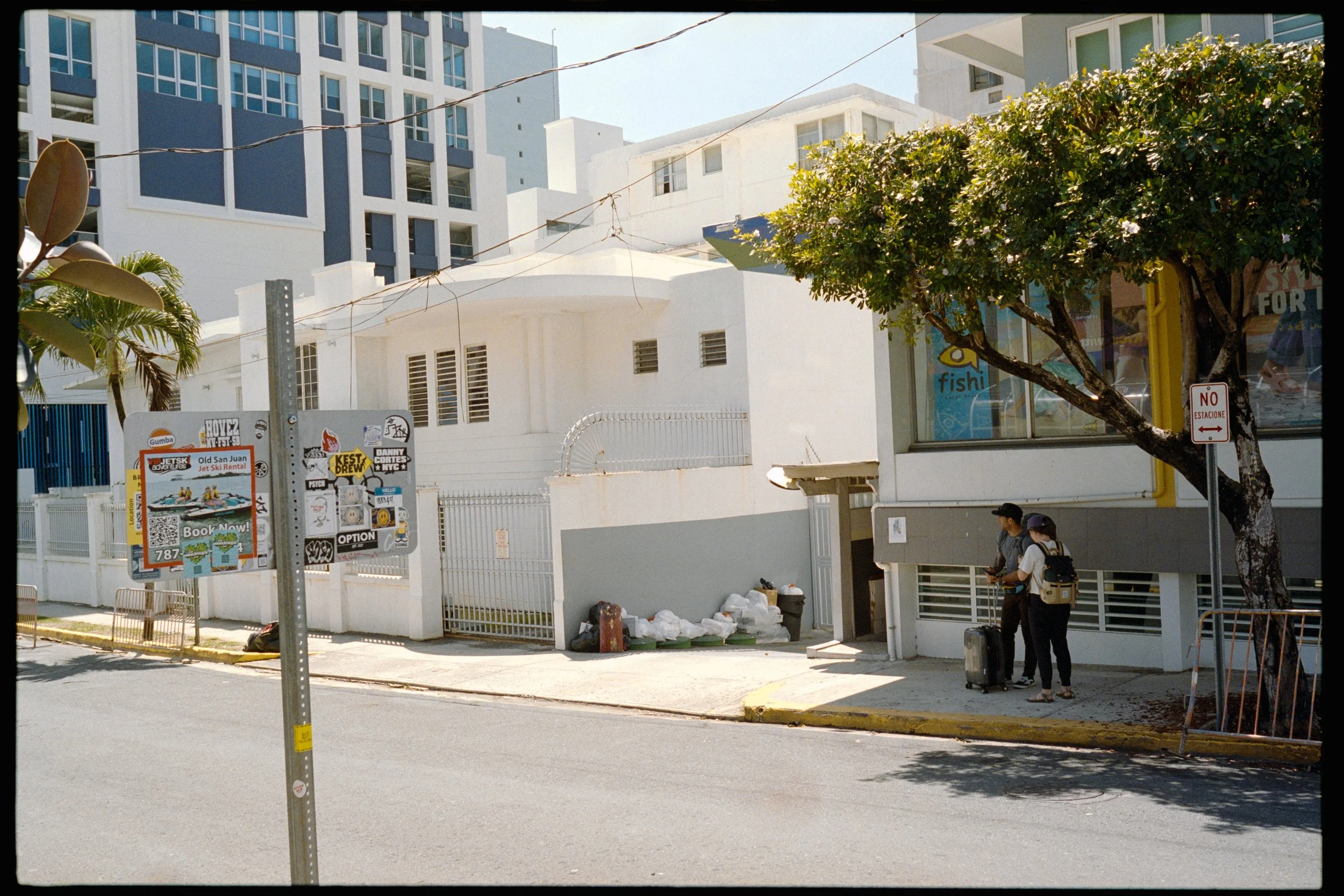 Street scene with modern white and gray buildings, a tree with green leaves, posters on a fence, and two people standing with luggage in front of a building.
