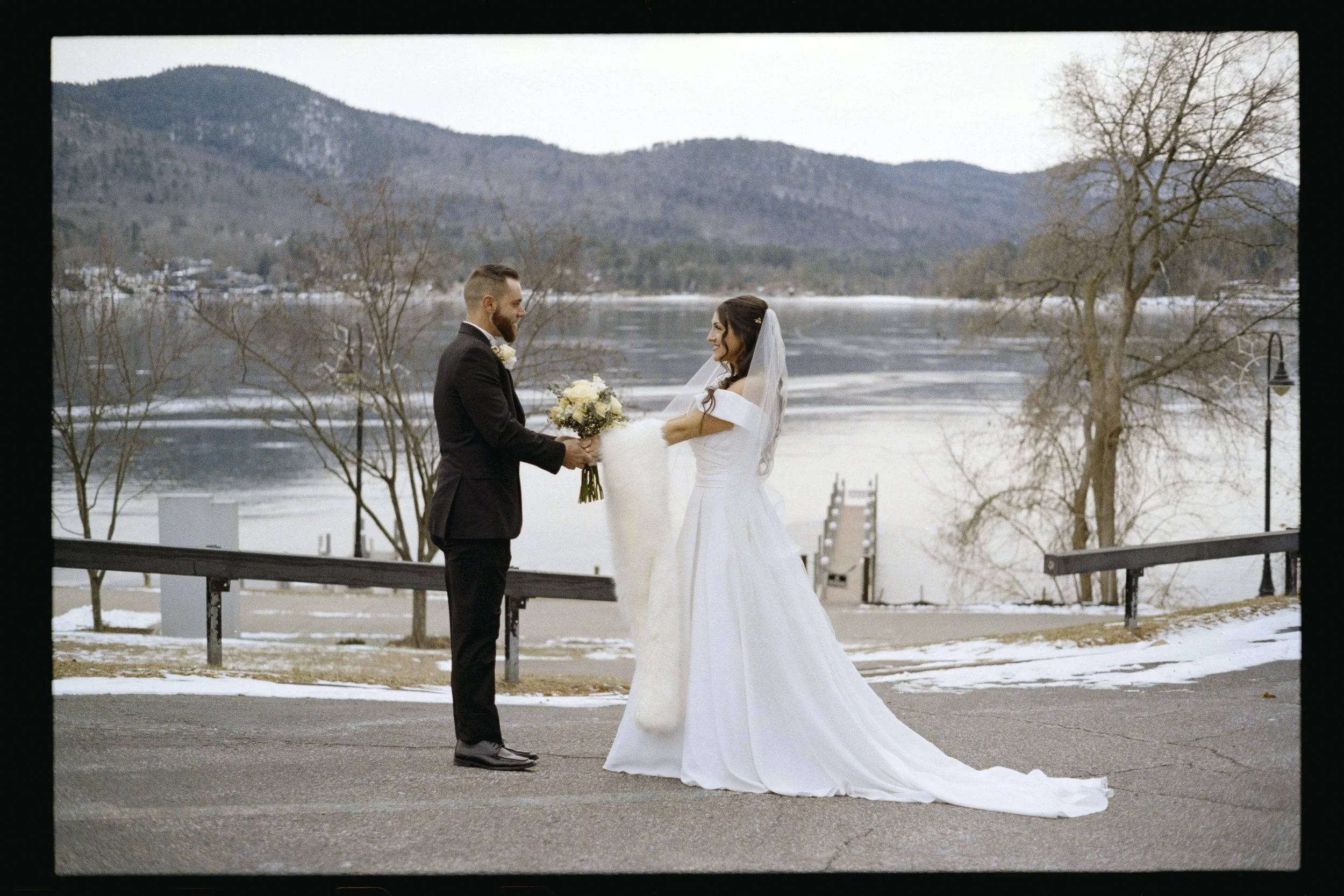 A bride and groom standing outdoors by a river, exchanging vows during a winter wedding. The groom is in a black tuxedo, and the bride is in a white wedding gown with a veil. Mountains, leafless trees, and snow are visible in the background.