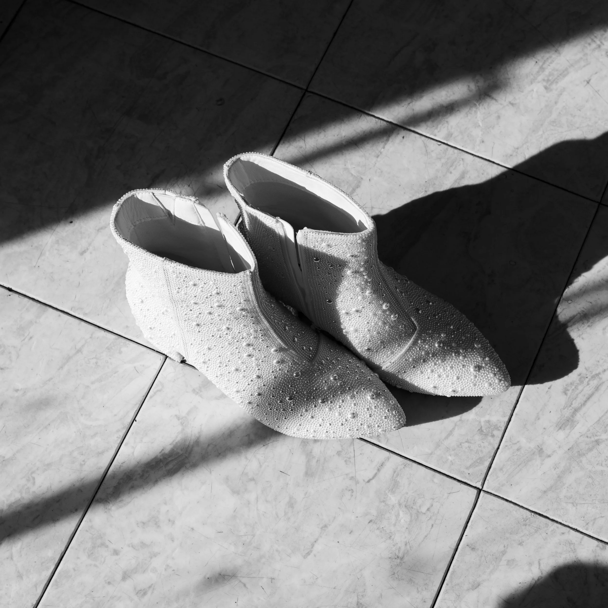 A pair of white sneakers with water droplets on them sitting on a tiled floor, with sunlight creating shadows.
