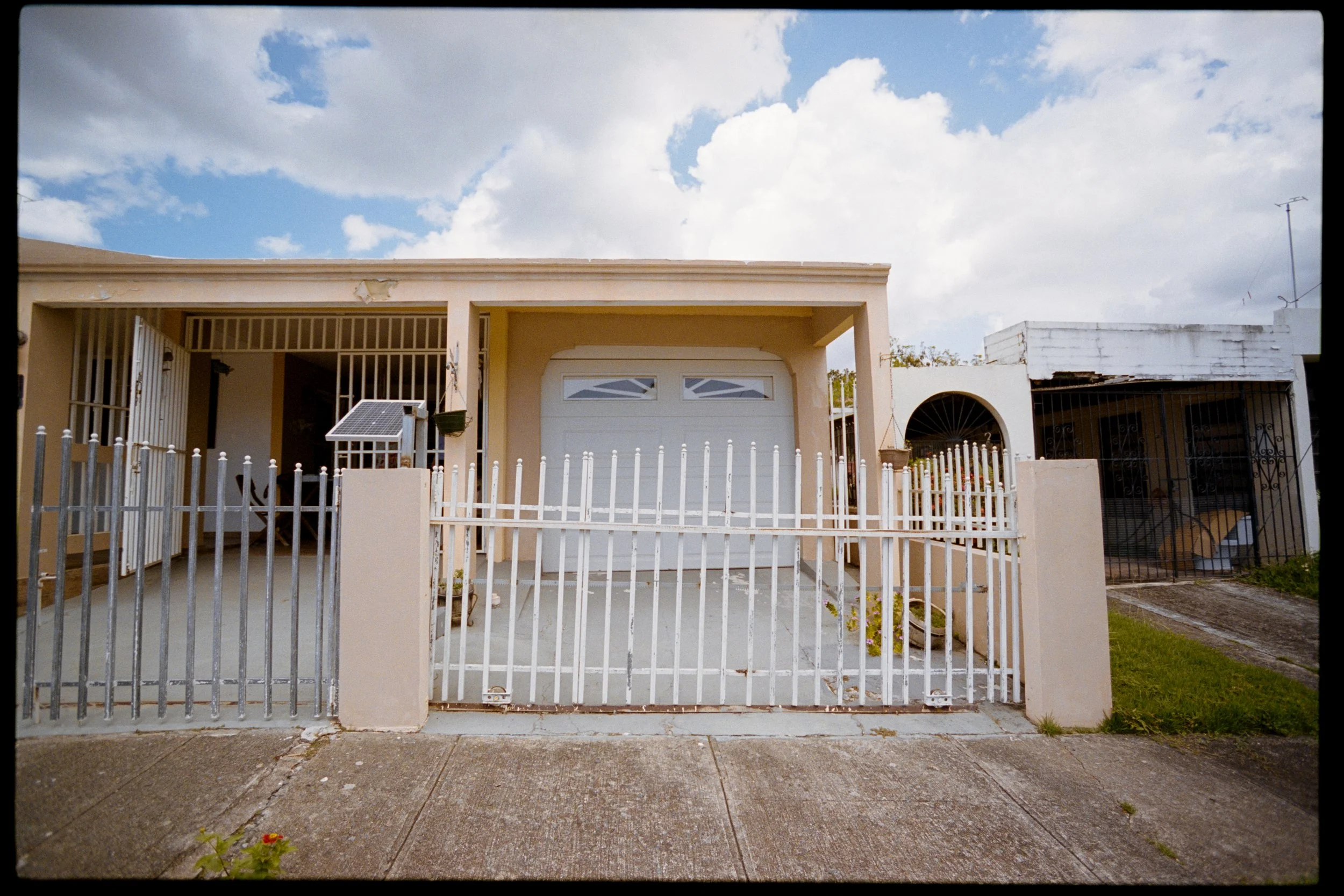 The front of a house with a white garage door under a beige porch, a metal fence, a solar panel on a post, and a sidewalk with a small plant growing through a crack. The sky is partly cloudy with patches of blue.