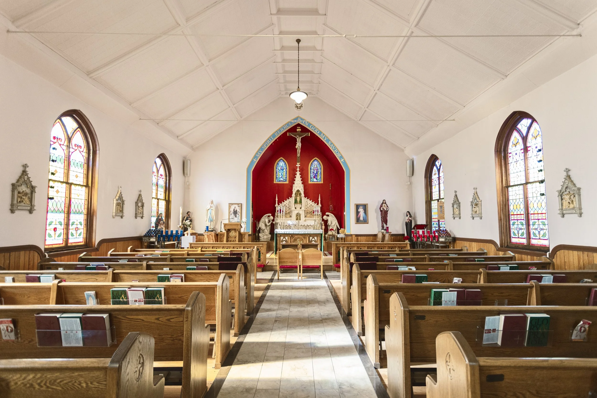 Interior of a church with wooden pews, stained glass windows, statues, and altar with religious symbols.