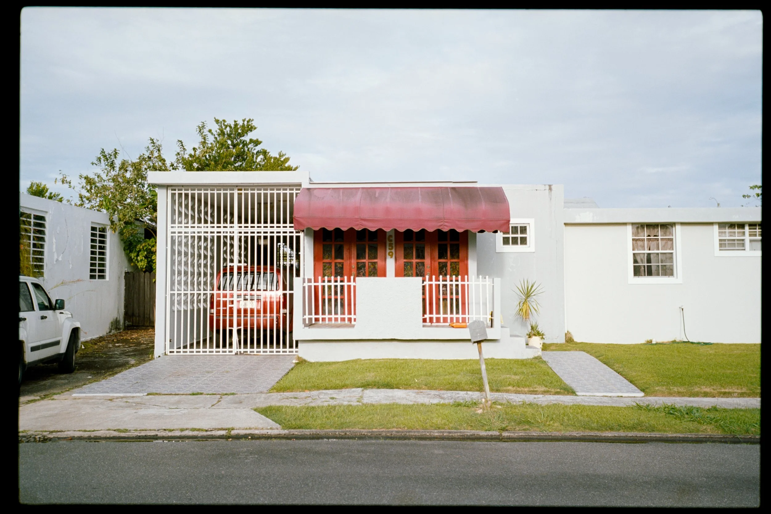 A single-story white house with a red awning over the front door, metal gate, and a small front yard with a grass lawn and paved walkway, parked cars on the street, and a cloudy sky overhead.