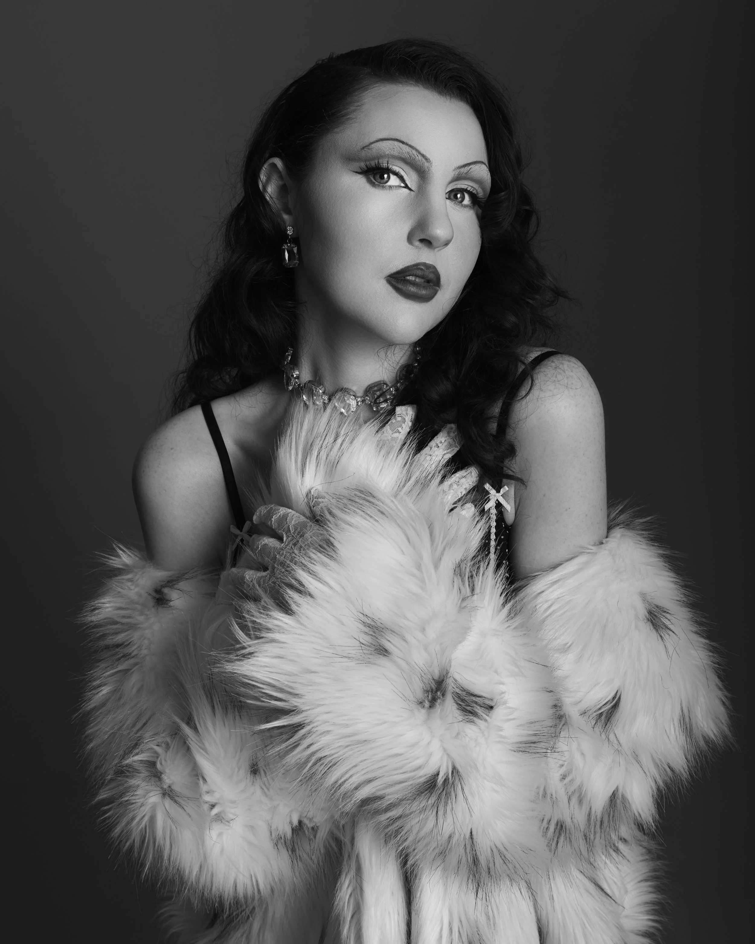 Black and white portrait of a woman with wavy dark hair, wearing makeup, jewelry, and a fur coat, against a plain dark background.