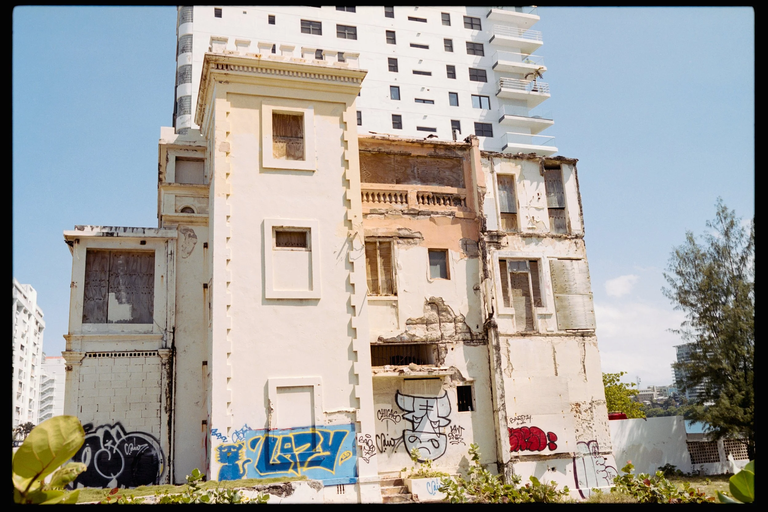 A partially demolished multi-story building with graffiti on its lower walls, with a modern white high-rise building visible in the background and a clear blue sky overhead.