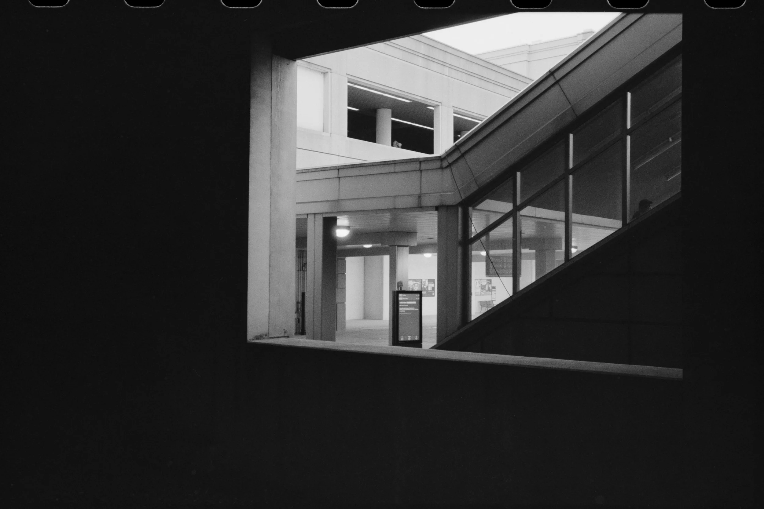 Black and white photo of an indoor architectural space with stairs and a covered balcony area. A person is visible on the upper level, looking through a window, and there is a digital information display on the lower level.