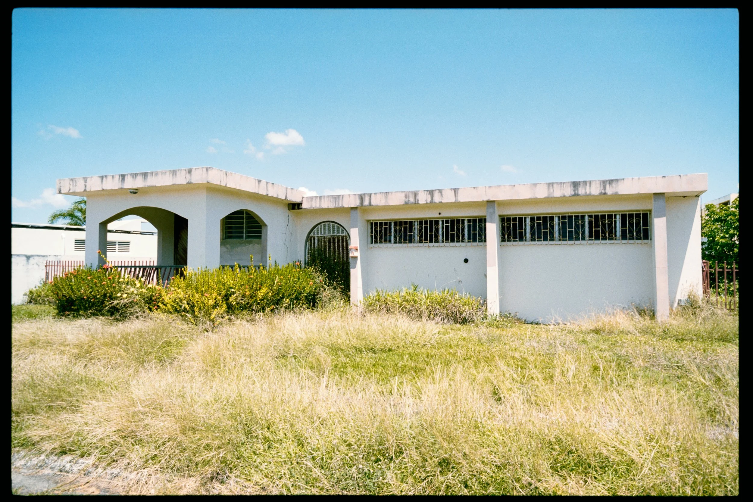 A single-story white house with a flat roof, a decorative archway entrance, and barred windows, surrounded by overgrown grass and bushes, under a bright blue sky with a few scattered clouds.