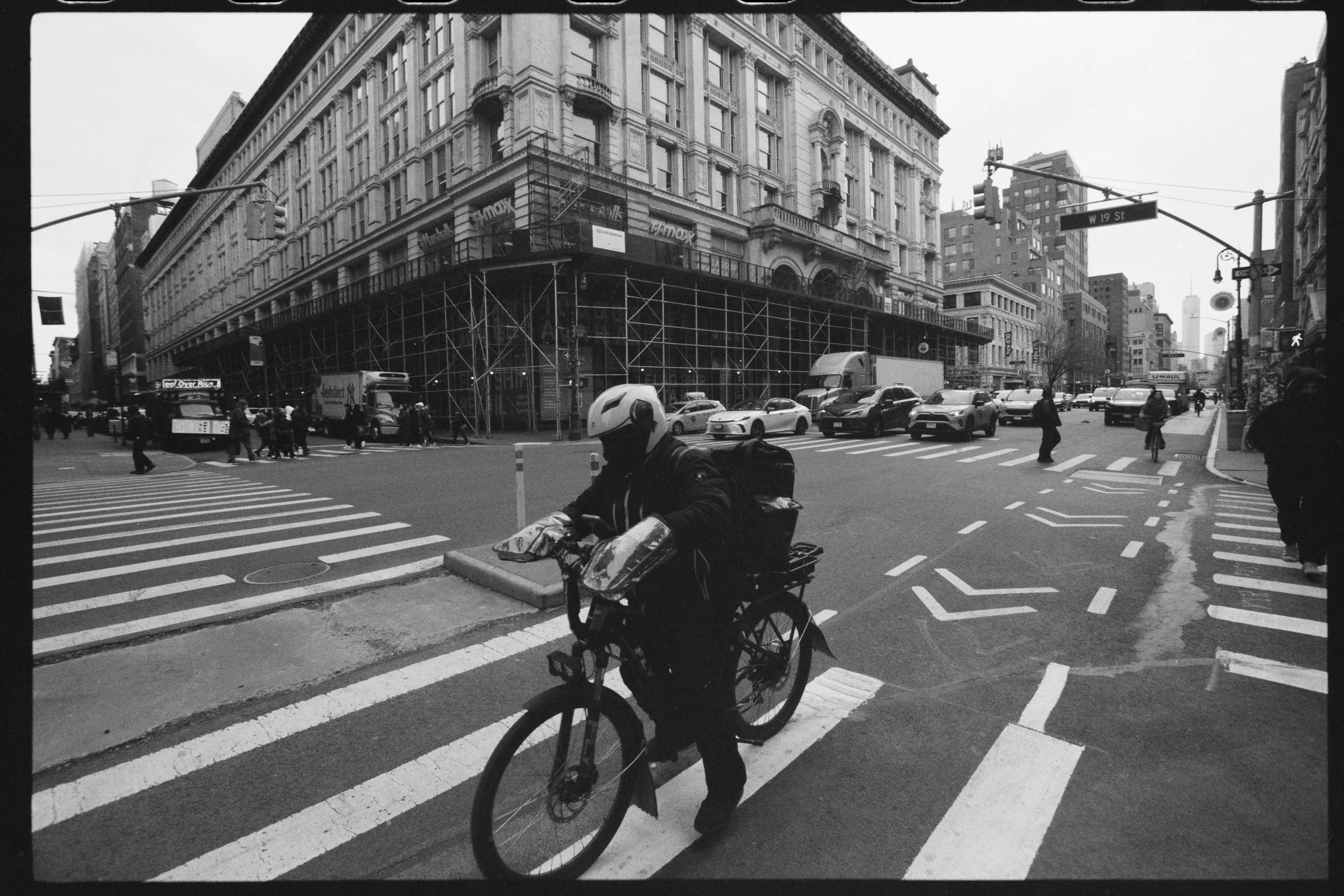 A black and white photo of a city street corner showing a large, ornate building under renovation with scaffolding. Pedestrians cross the crosswalk and a cyclist is riding through the intersection. There are various vehicles on the street including c