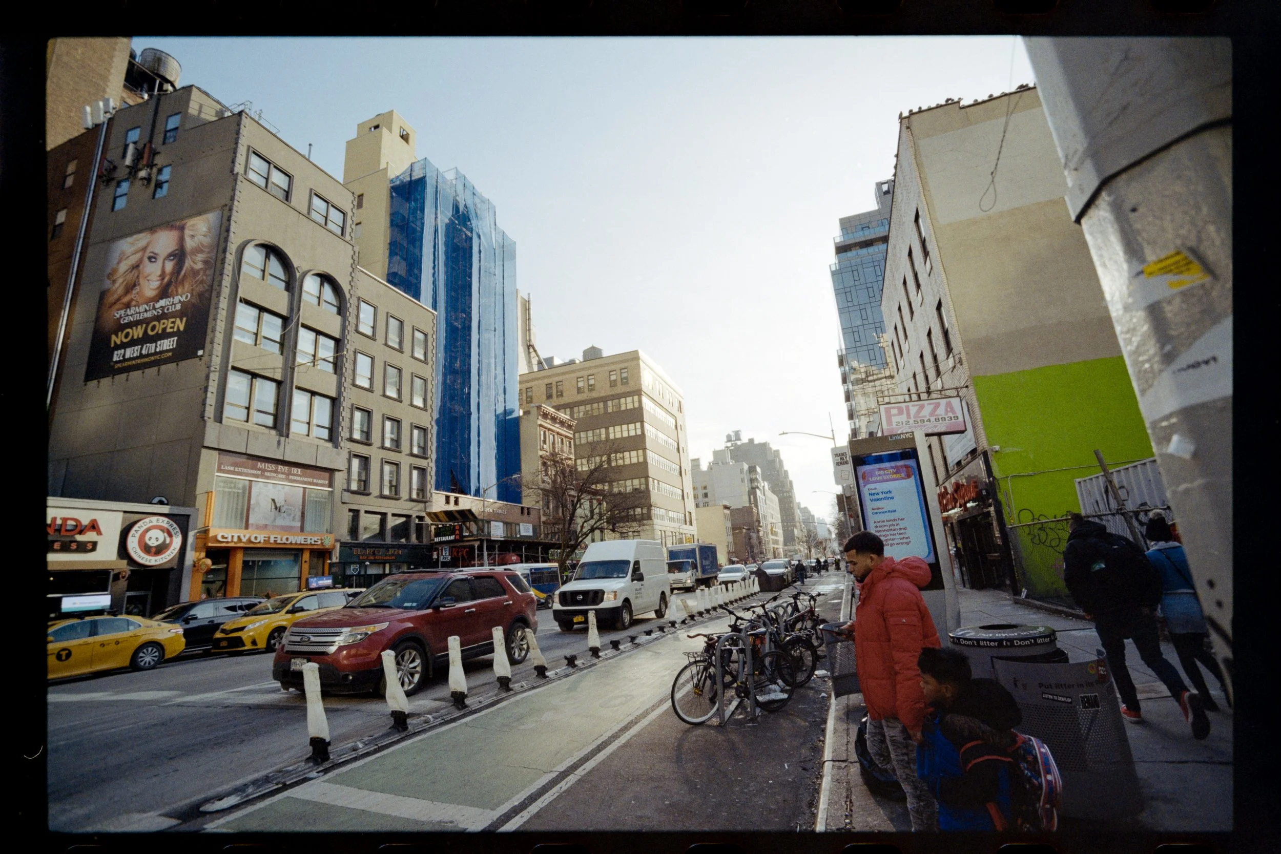 City street scene with parked cars, bikes, pedestrians, and tall buildings, including billboards and construction scaffolding.