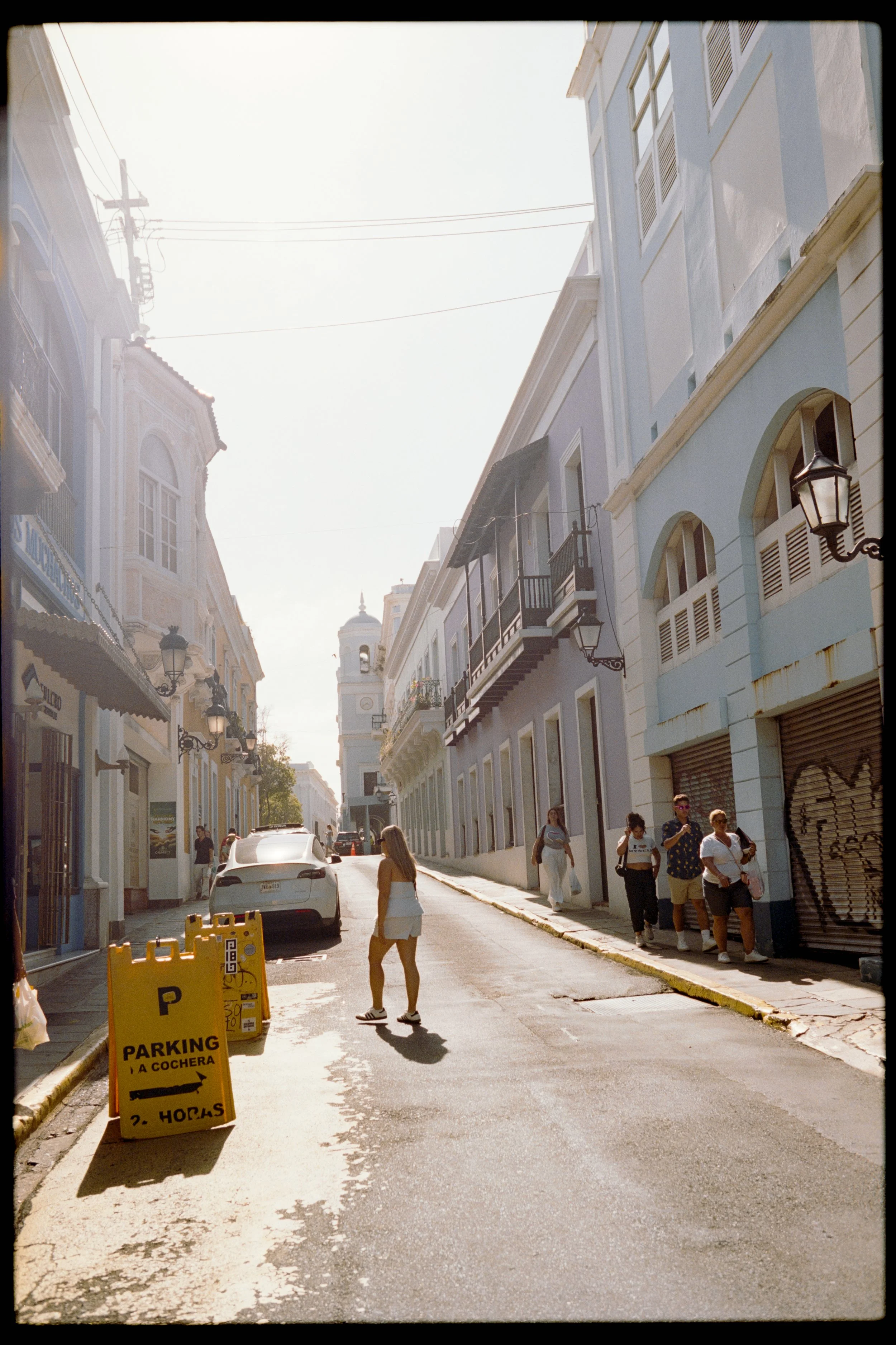 A street scene with pastel-colored buildings, pedestrians walking, a parked white car, and a yellow parking sign in Spanish indicating parking and parking hours, with the bright sky overhead.