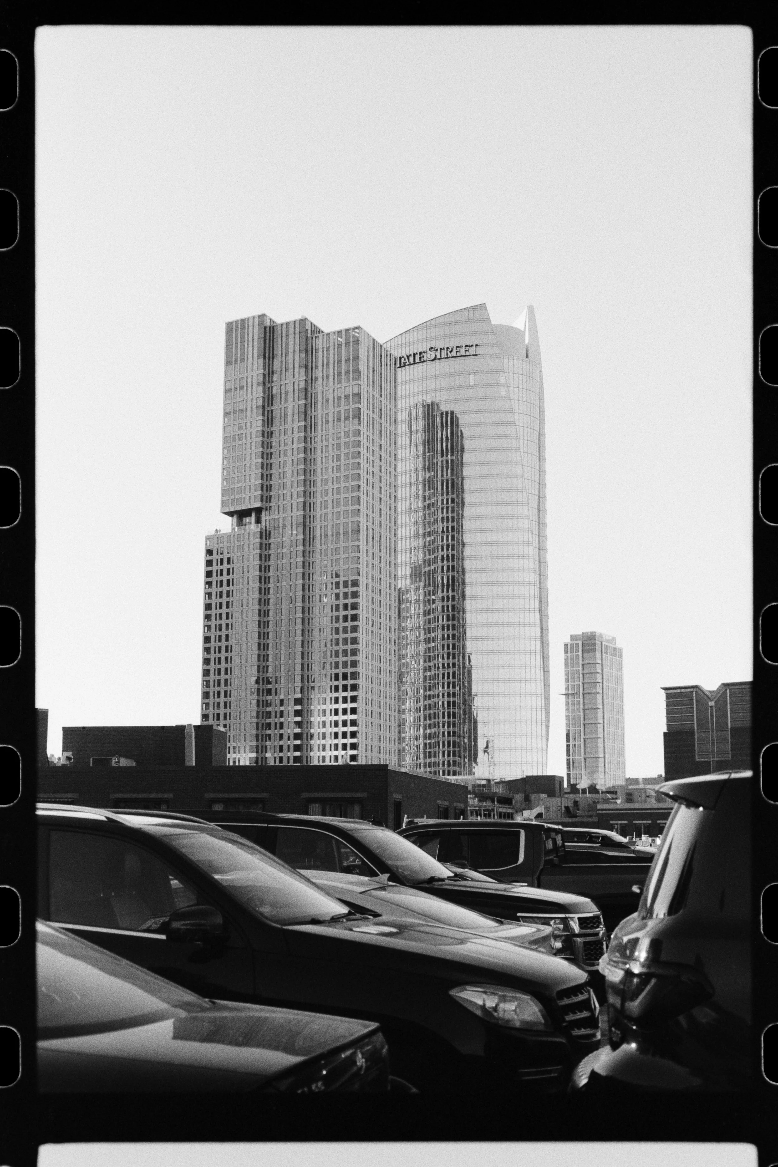Black and white photo of tall, modern skyscrapers with glass facades, with cars parked in a lot in the foreground.