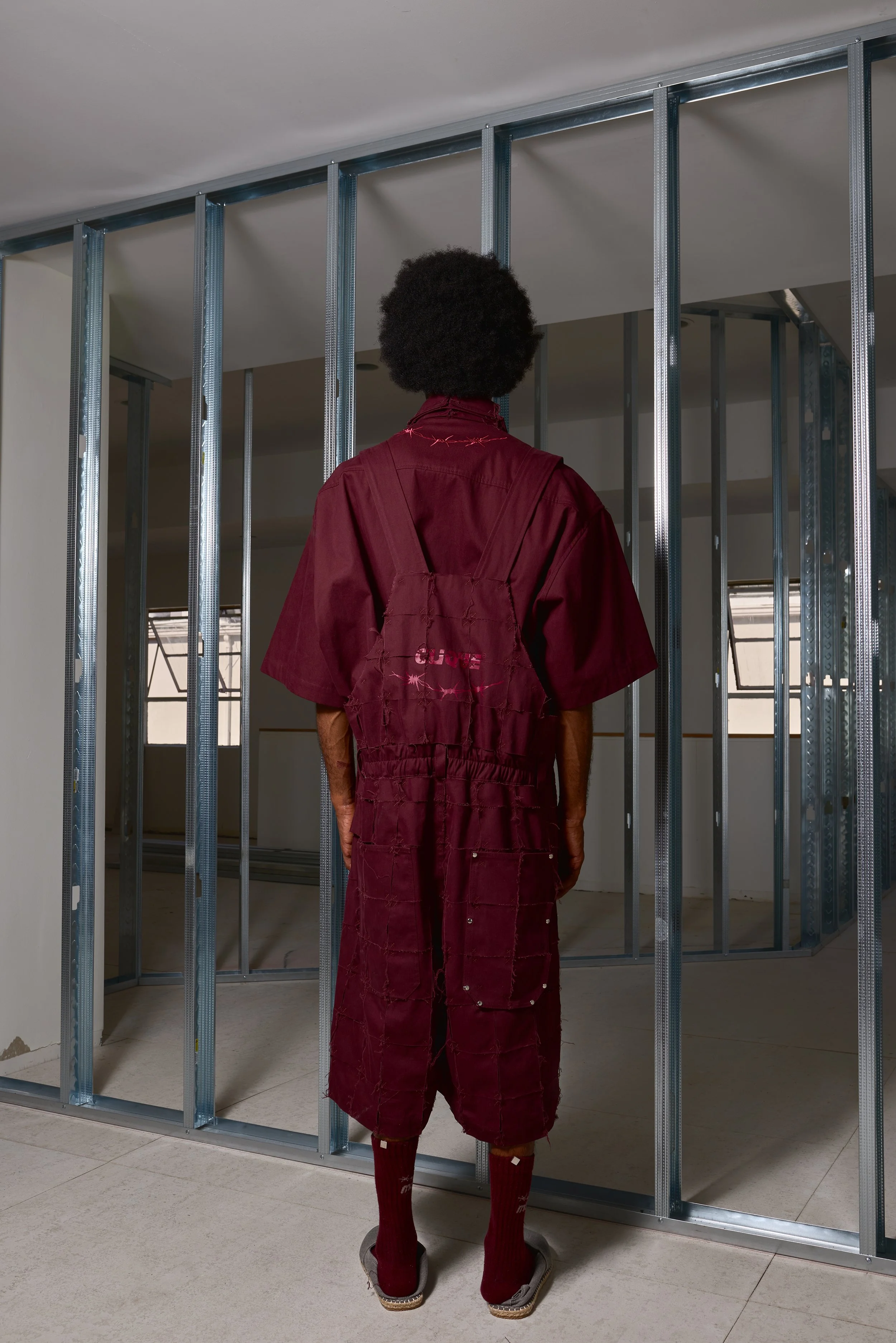 Person with an afro hairstyle in construction attire standing in front of a metal wall frame, wearing a burgundy oversized shirt and pants with patches, and maroon socks with sandals.