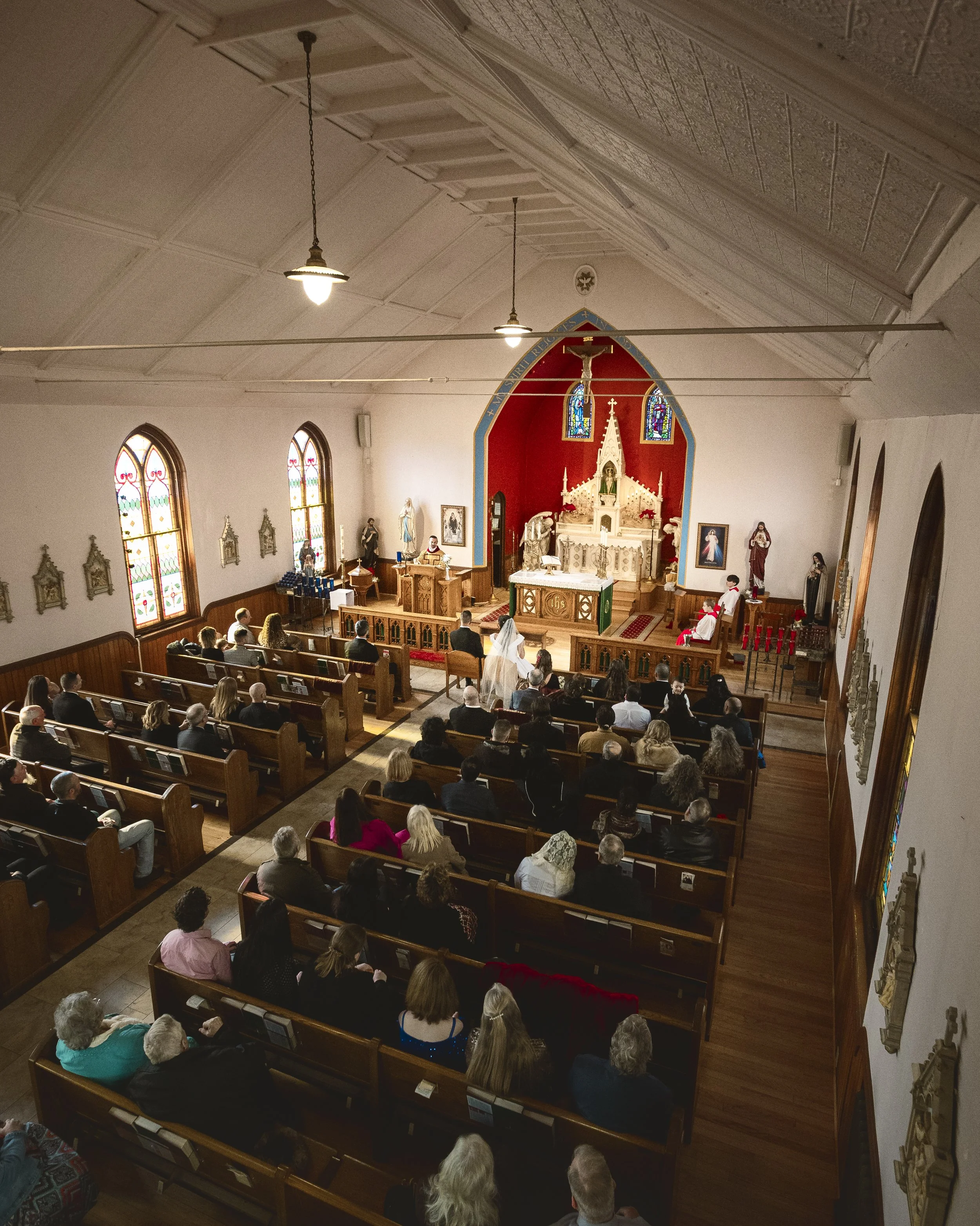 Interior of a church during a wedding ceremony, with pews filled with guests, an altar with a bride and groom, and stained glass windows.