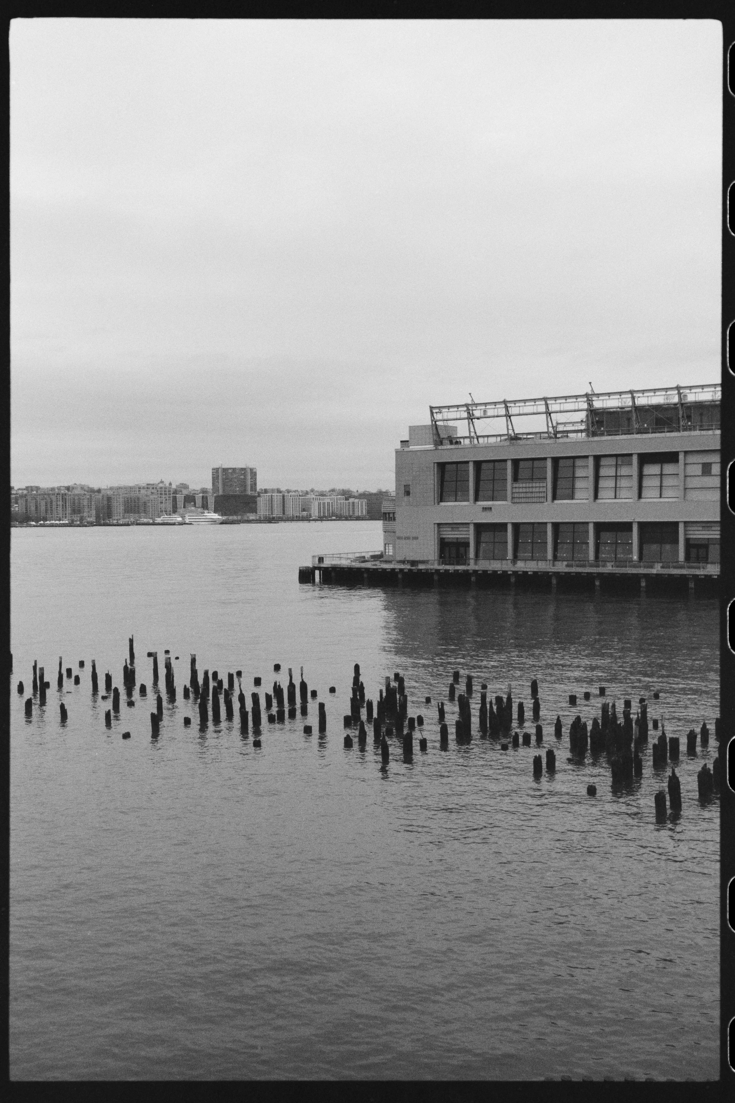 Black and white photo of a city skyline across a body of water, with buildings and a large rectangular structure on the water in the foreground. Wooden pilings in the water are also visible.