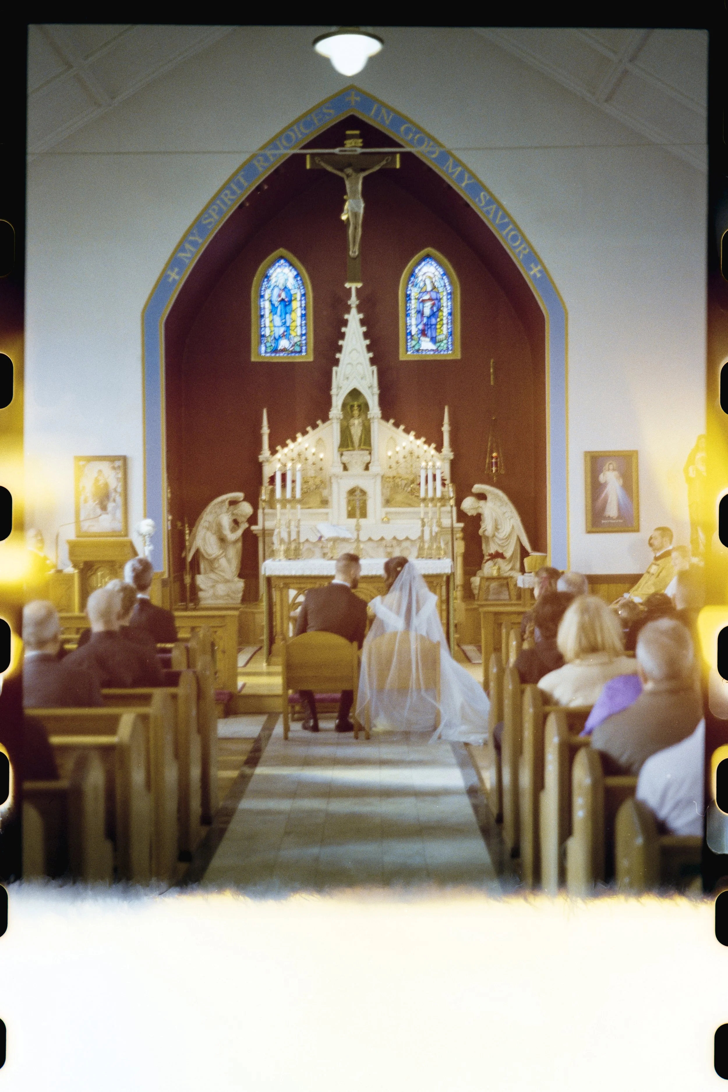Inside a church with an altar, crucifix, stained glass windows, and people attending a wedding ceremony.