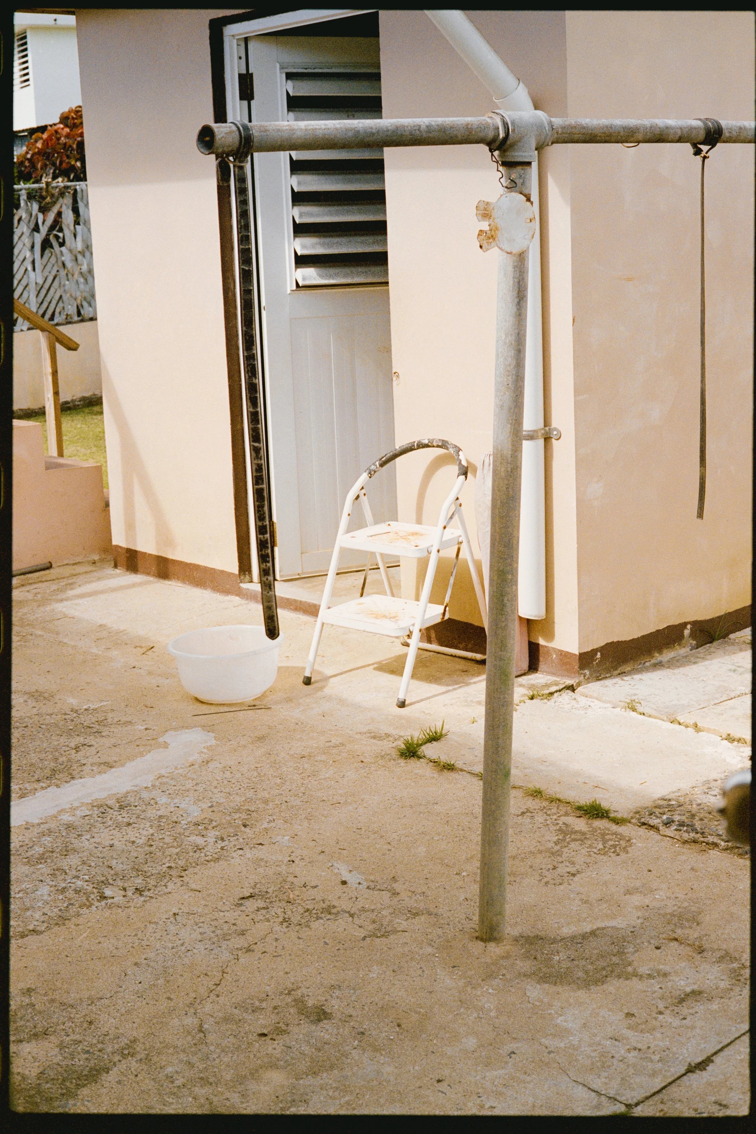 A white plastic chair and a plastic bucket on a cracked concrete ground outside a beige-colored building. There is a metallic stand with hooks, a small white pipe on the right, and an open vent on the building wall. A small tuft of grass is growing t