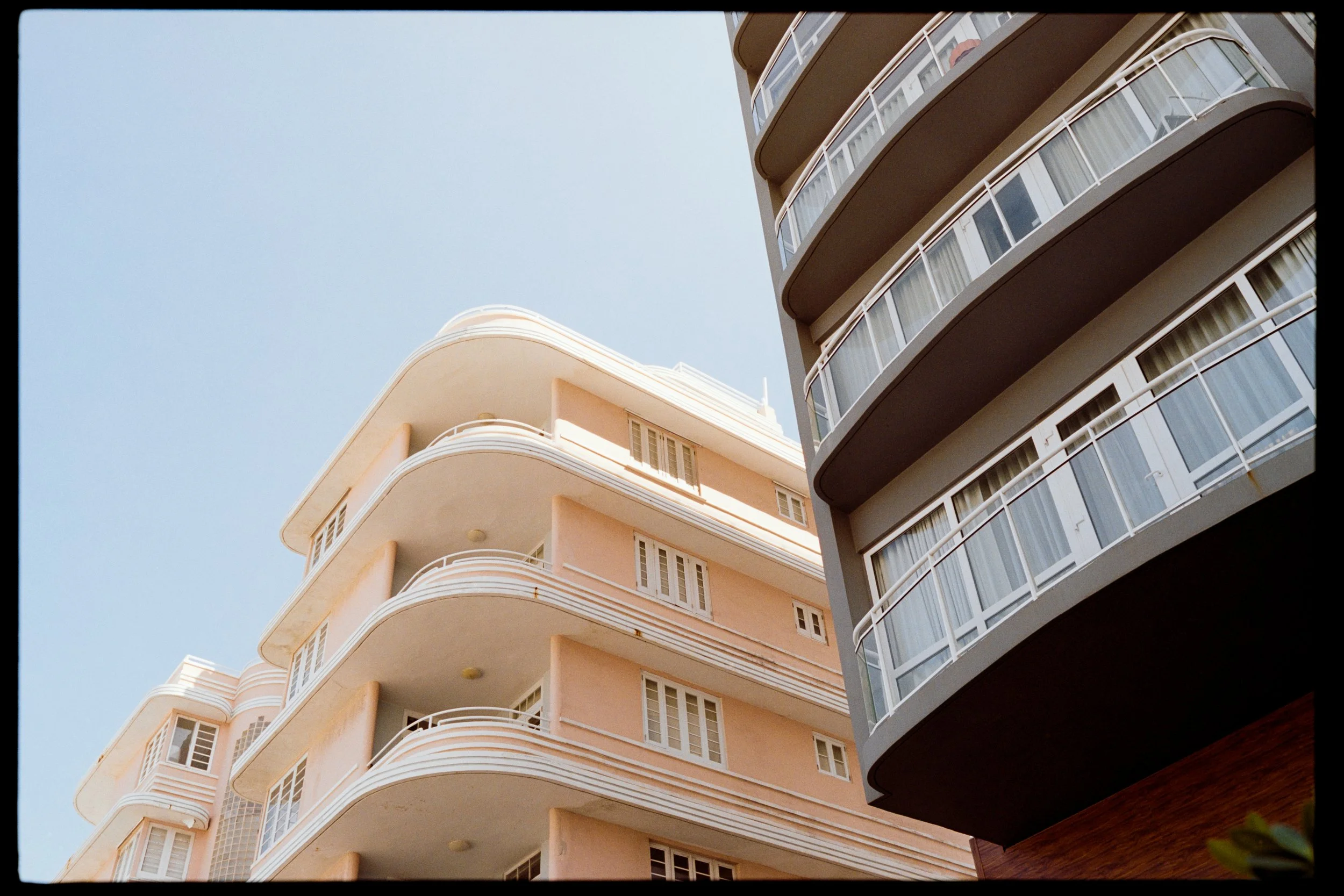 View of two modern high-rise apartment buildings with rounded balconies, against a clear blue sky. The building on the left is light pink and the building on the right is gray.