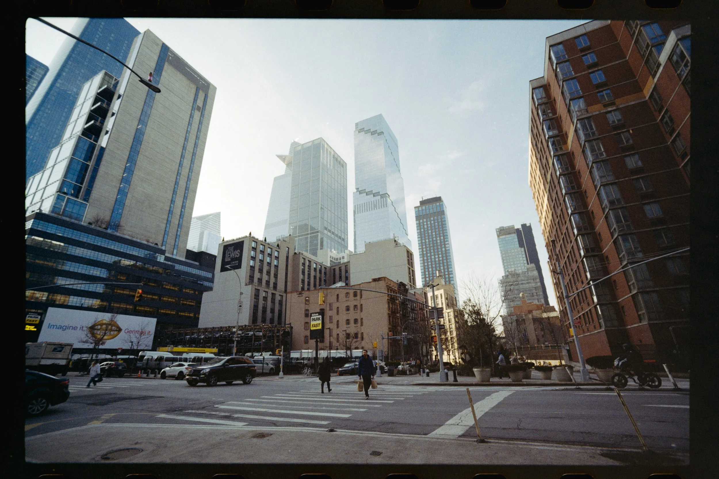 A city street scene with tall modern skyscrapers in the background, a busy crosswalk with pedestrians, and cars on the road. There are commercial buildings, billboards, and street signs visible. The sky is partly cloudy.
